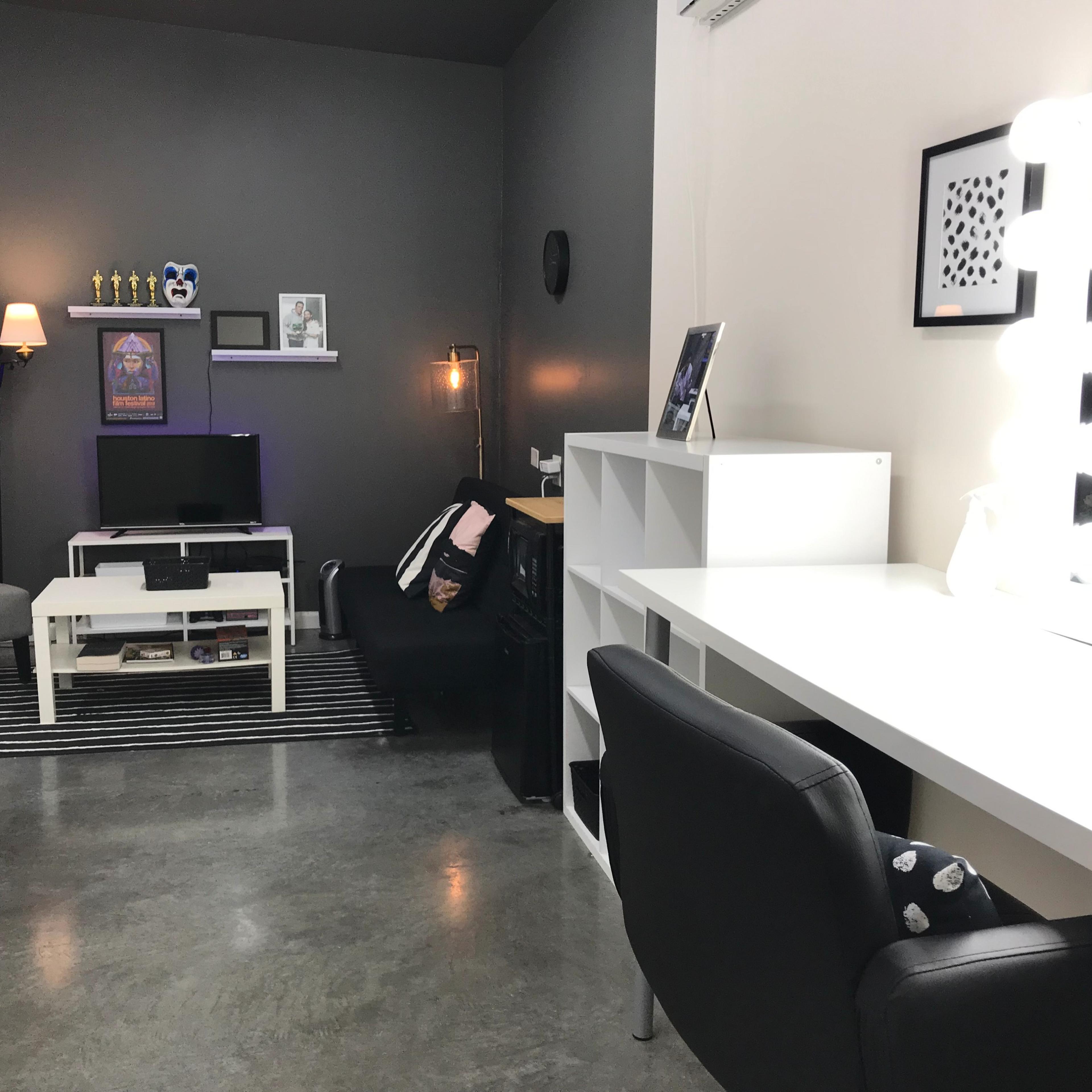 A modern room with a sitting area featuring a gray chair and a television, along with a vanity desk illuminated by a lighted mirror.
