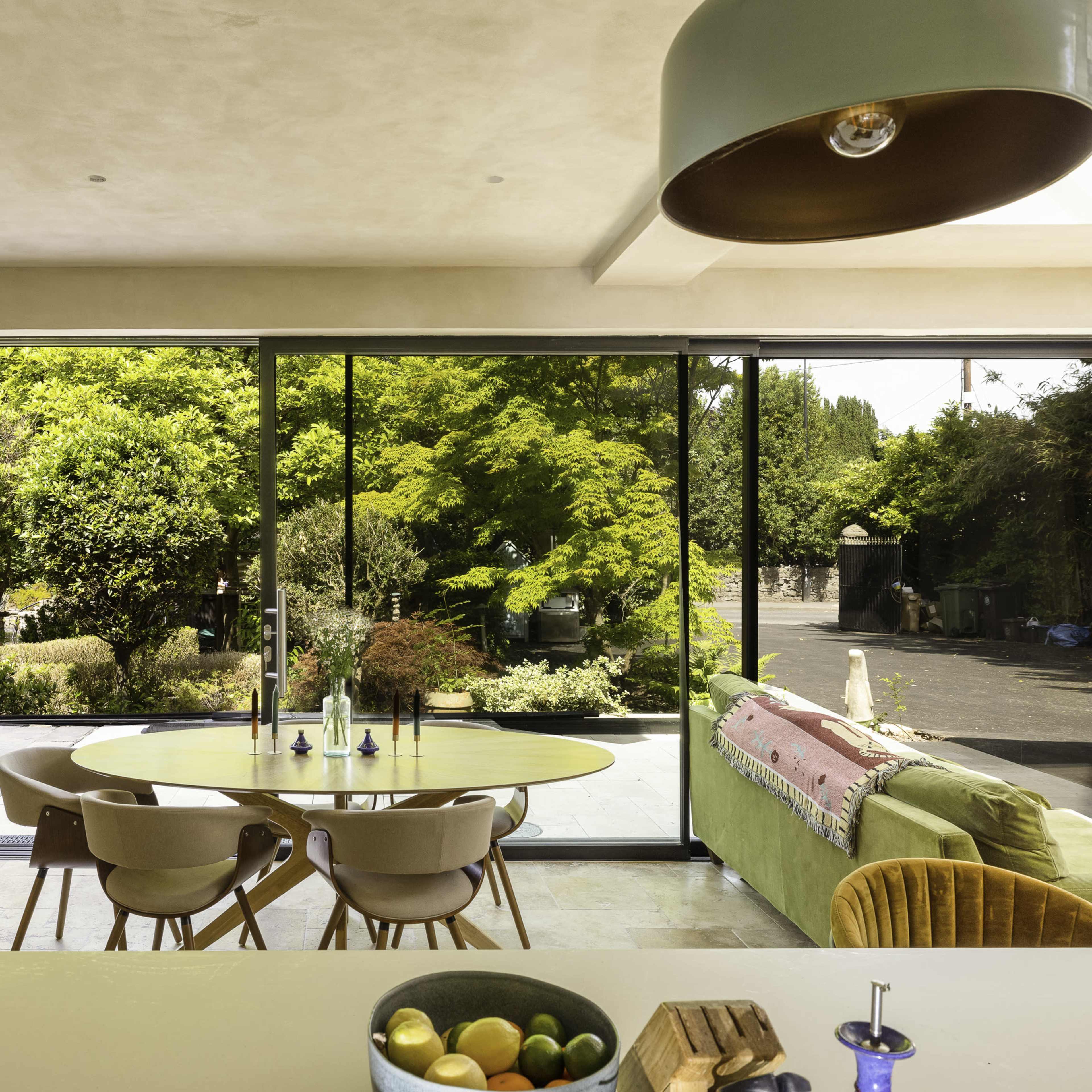 A modern kitchen area features a round dining table with chairs, surrounded by large windows that showcase a lush garden outside.