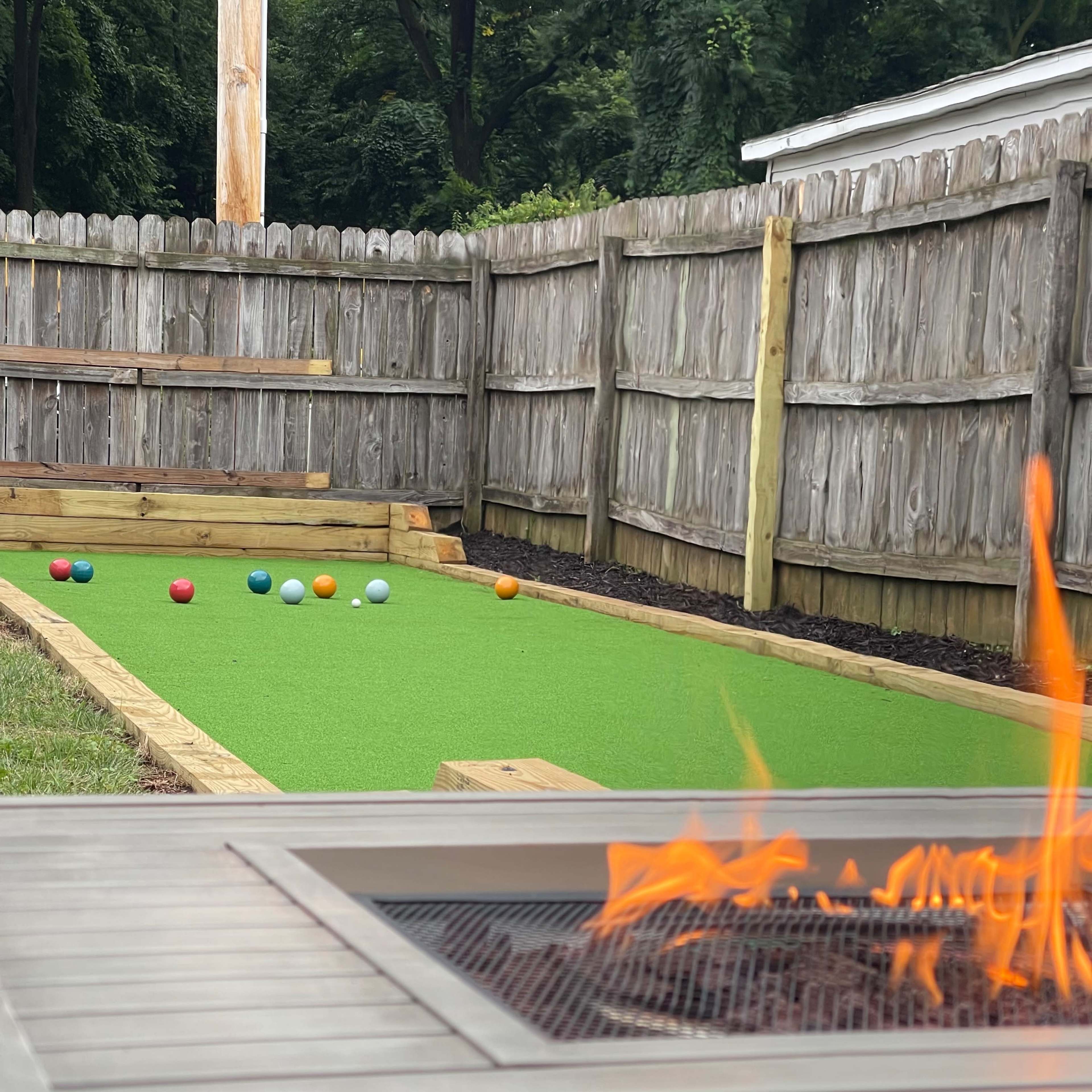A backyard with a bowling green, featuring colored balls arranged on a synthetic grass surface and a fire pit in the foreground.