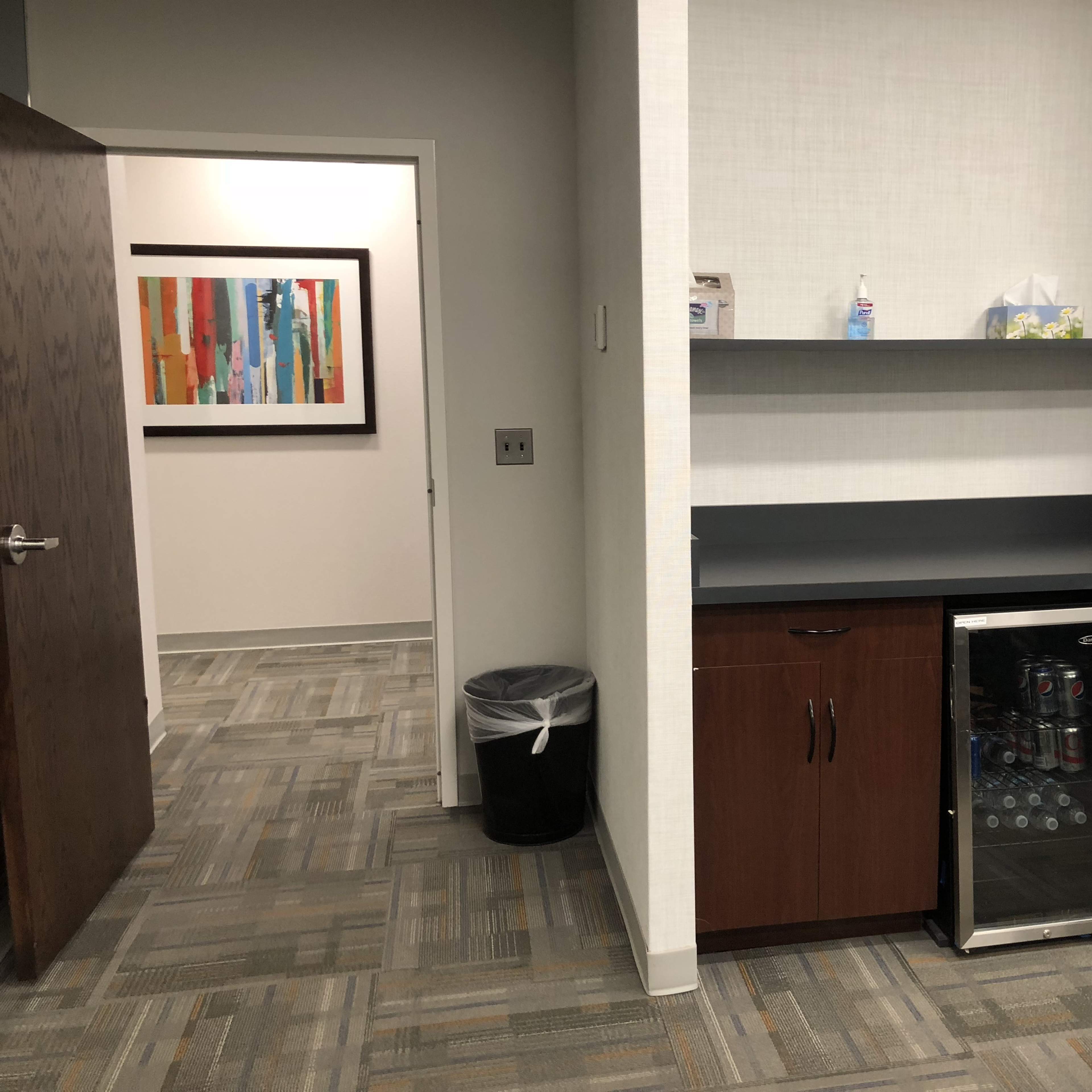 A small kitchenette area with a mini-fridge, a countertop, and a trash can, adjacent to an open door leading into another room.