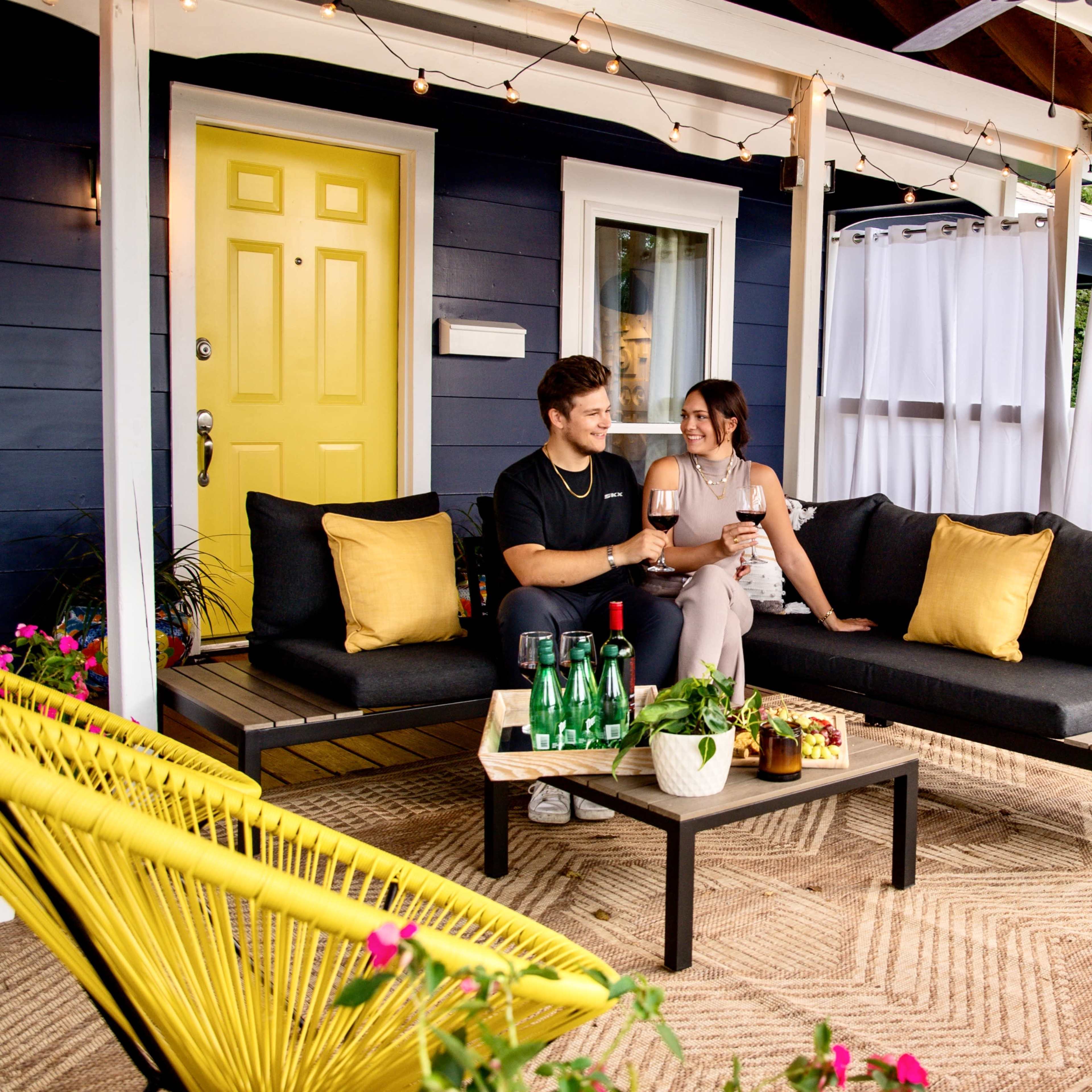 A couple sits on a patio with black couches and yellow pillows, enjoying drinks at a table on a deck decorated with plants and white curtains.