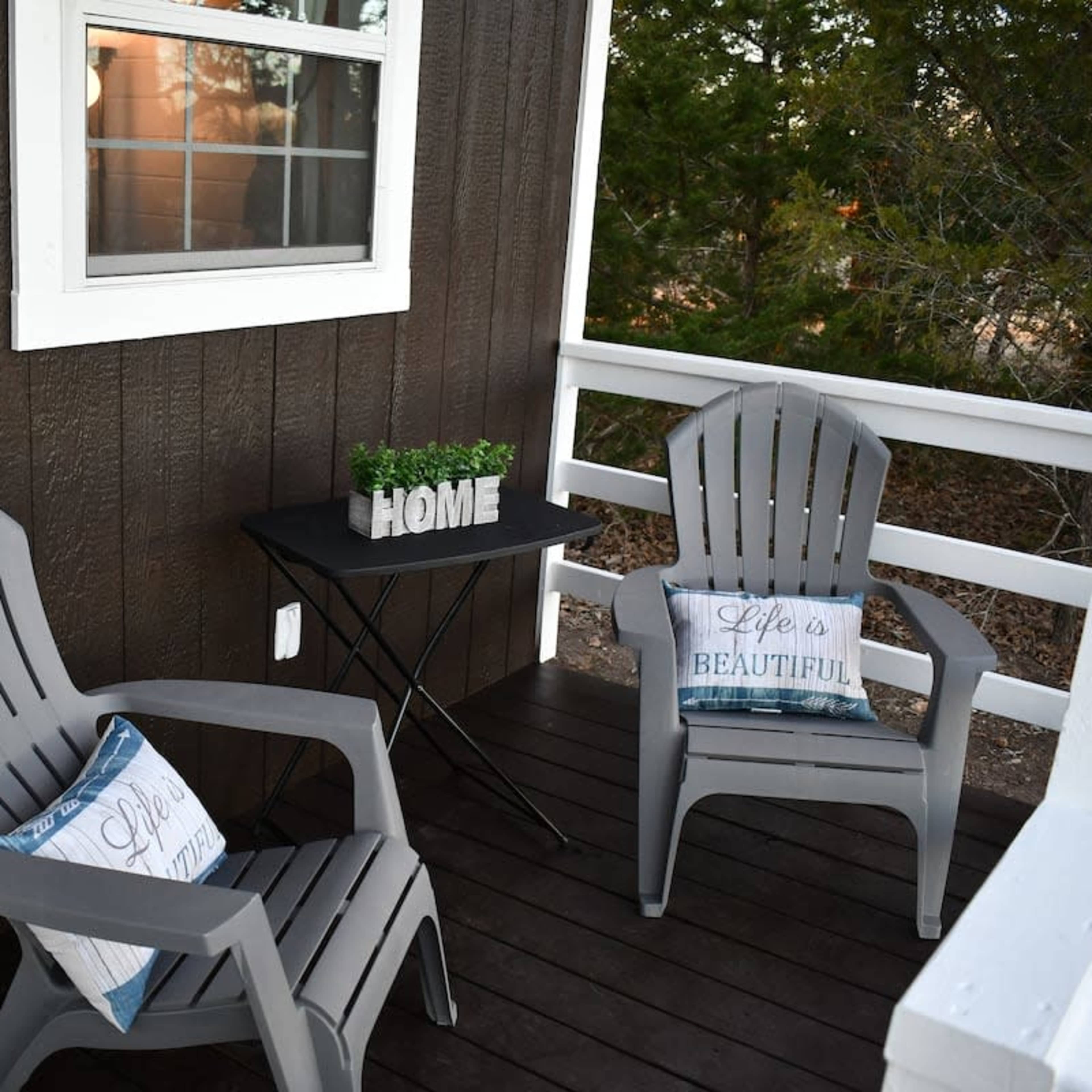 The scene features two gray outdoor chairs facing a small table with the word "HOME" and a potted plant, set on a dark wooden porch surrounded by greenery.