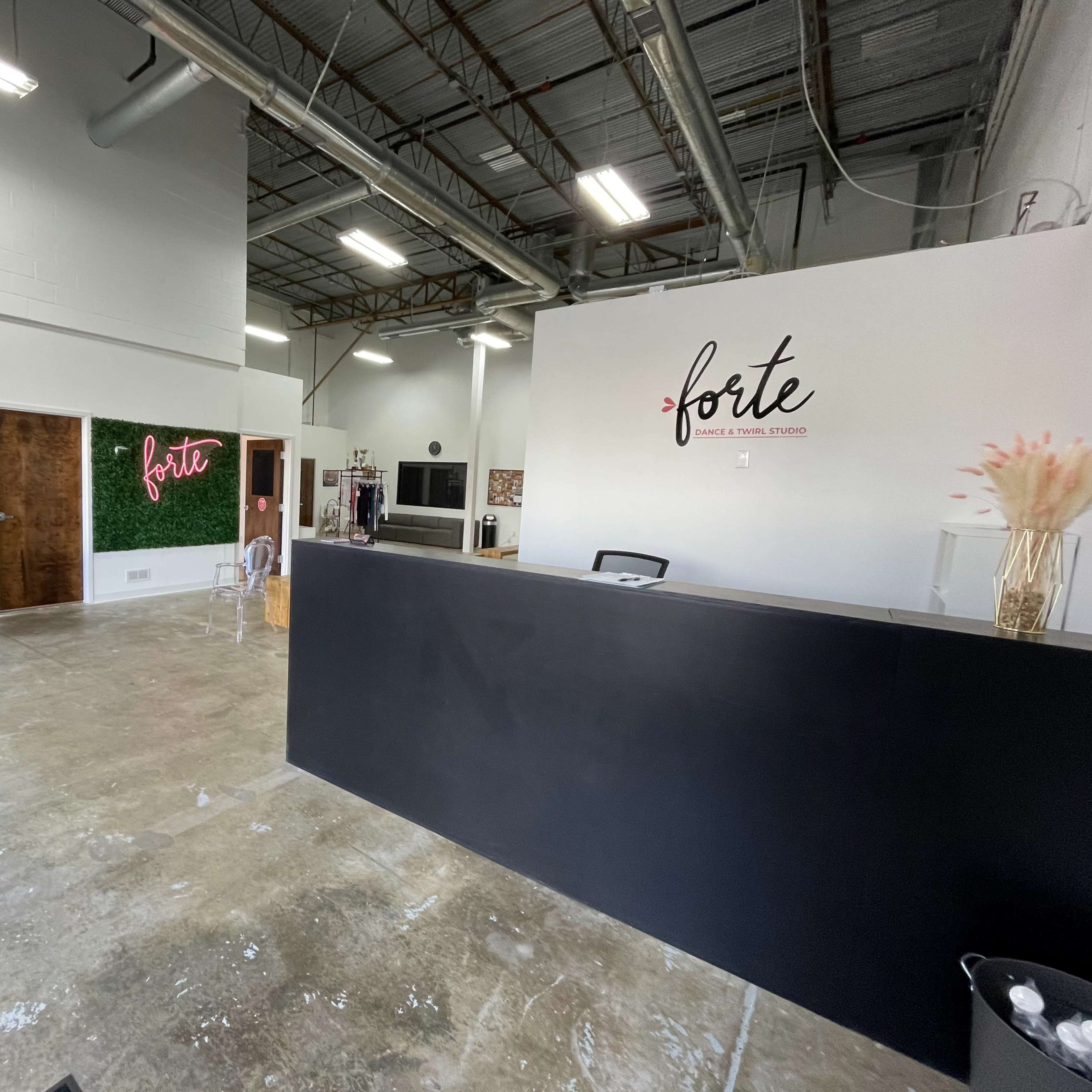 A modern interior of a studio or office space with a reception area featuring a black counter and decorative wall signage, alongside a seating area with white chairs and a green plant wall.