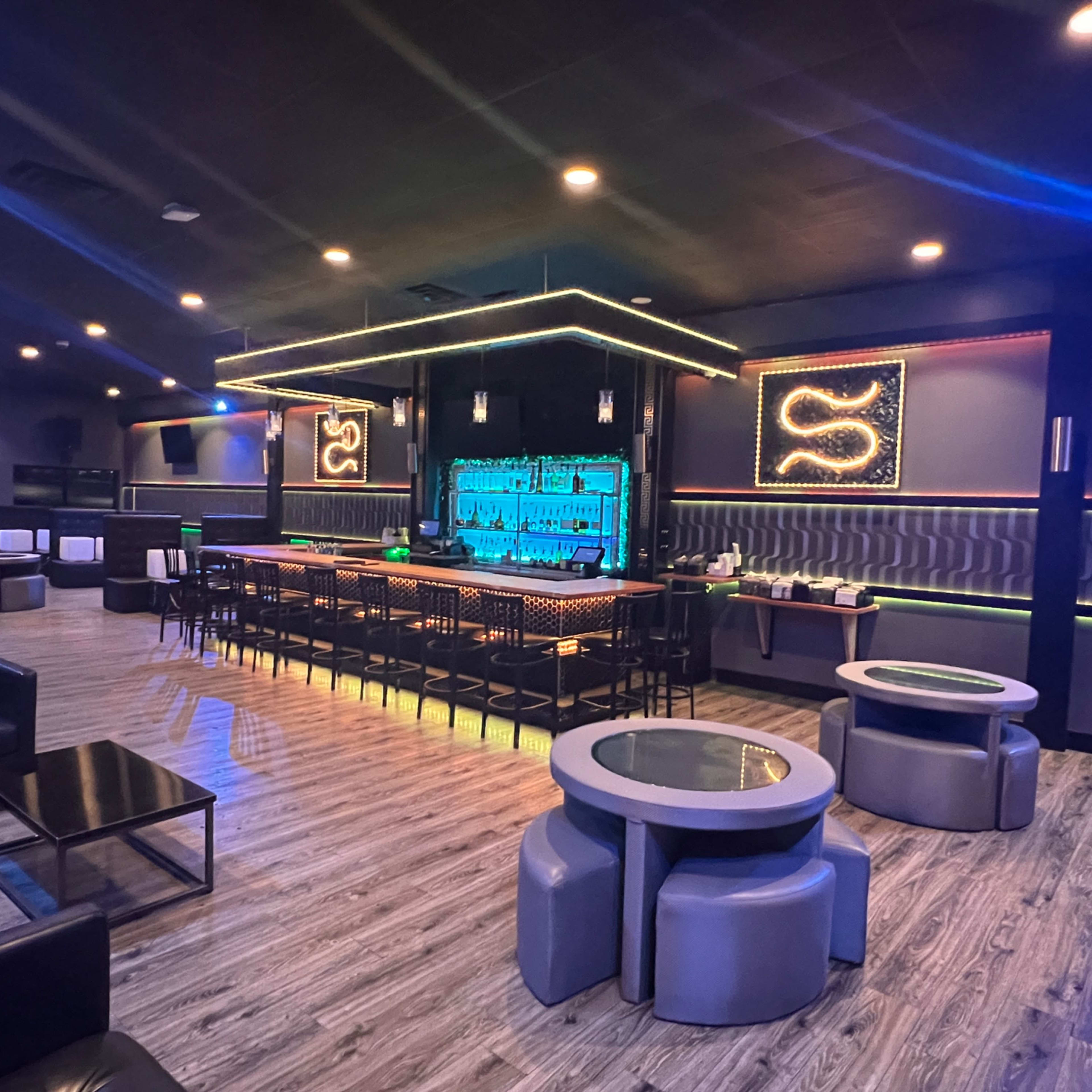 A modern lounge interior with a sleek bar, illuminated by blue lighting, surrounded by black leather seating and circular tables.