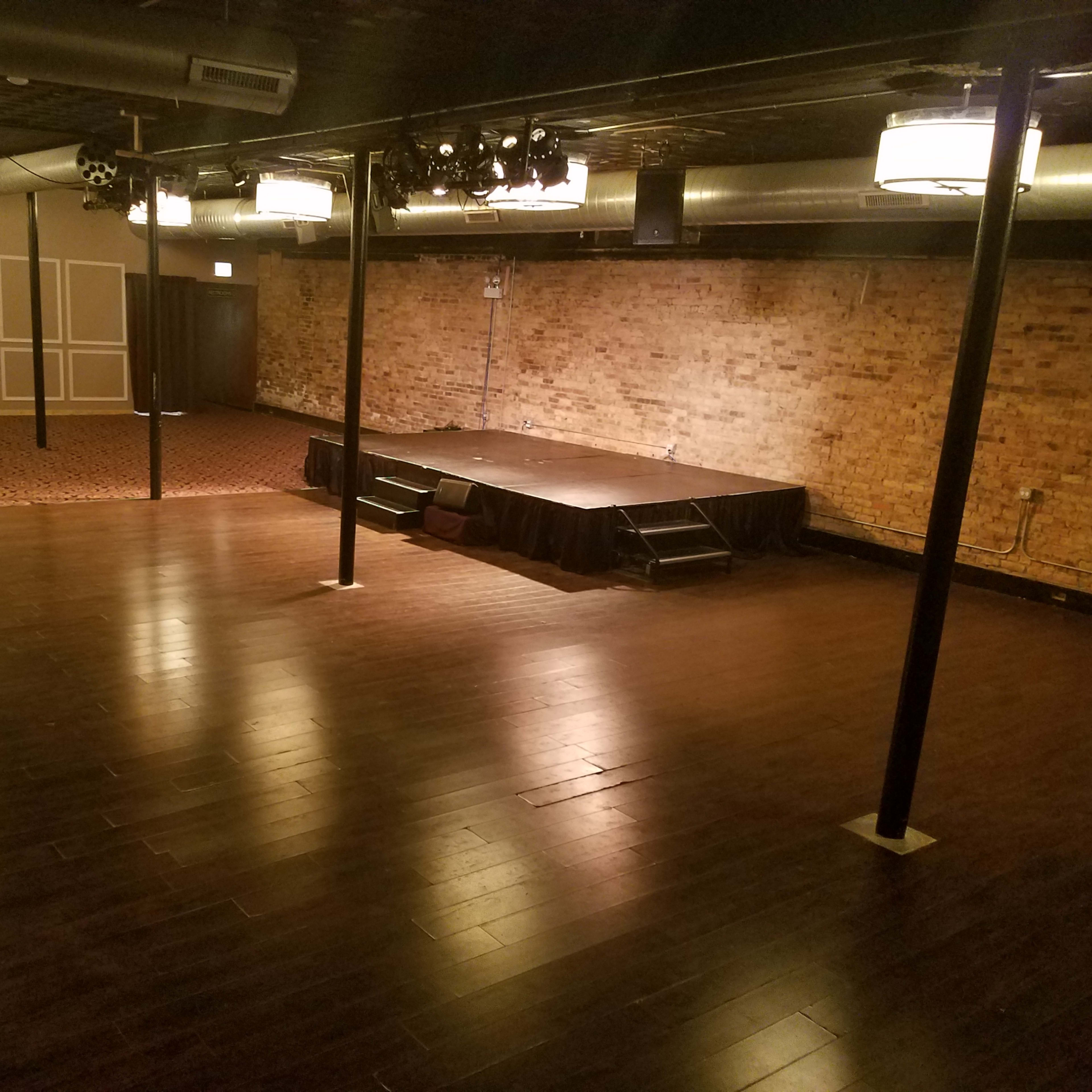 A spacious, empty event venue with wooden floors, a stage at the far end, and exposed brick walls.