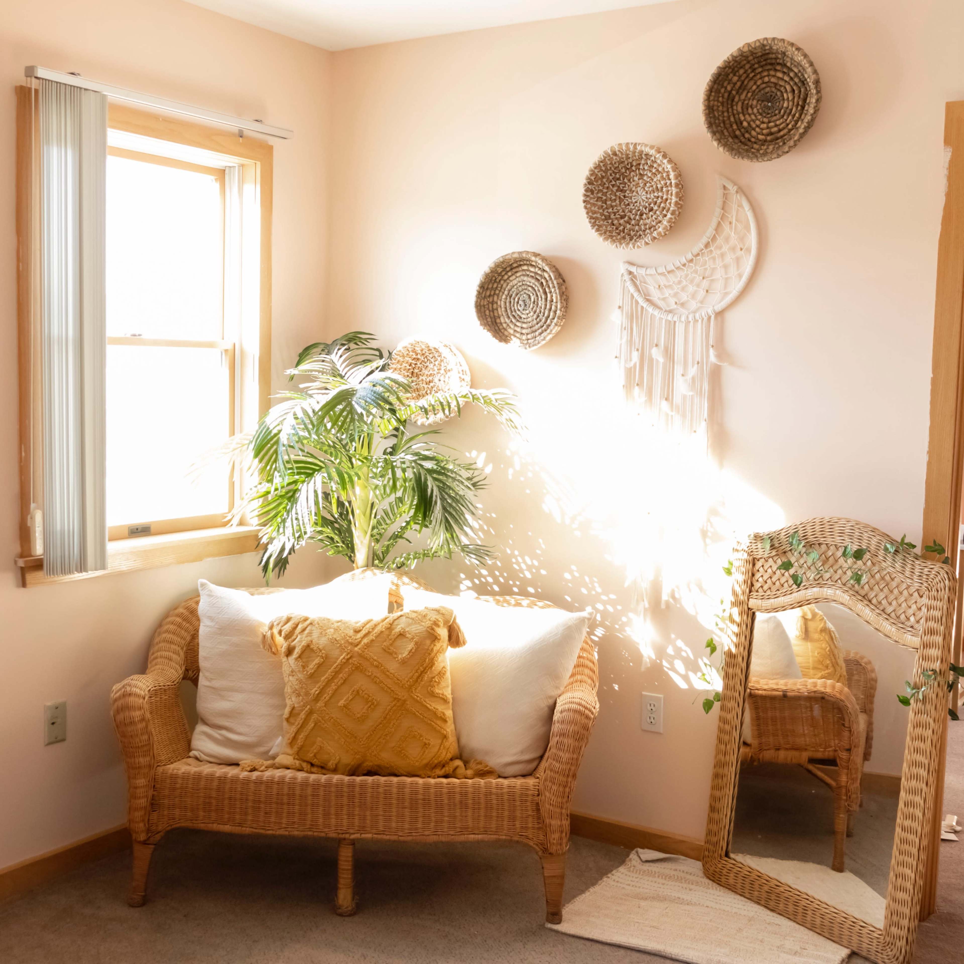 A cozy corner of a room features a rattan loveseat with decorative pillows, a potted plant, and wall hangings, illuminated by natural light streaming through a window.