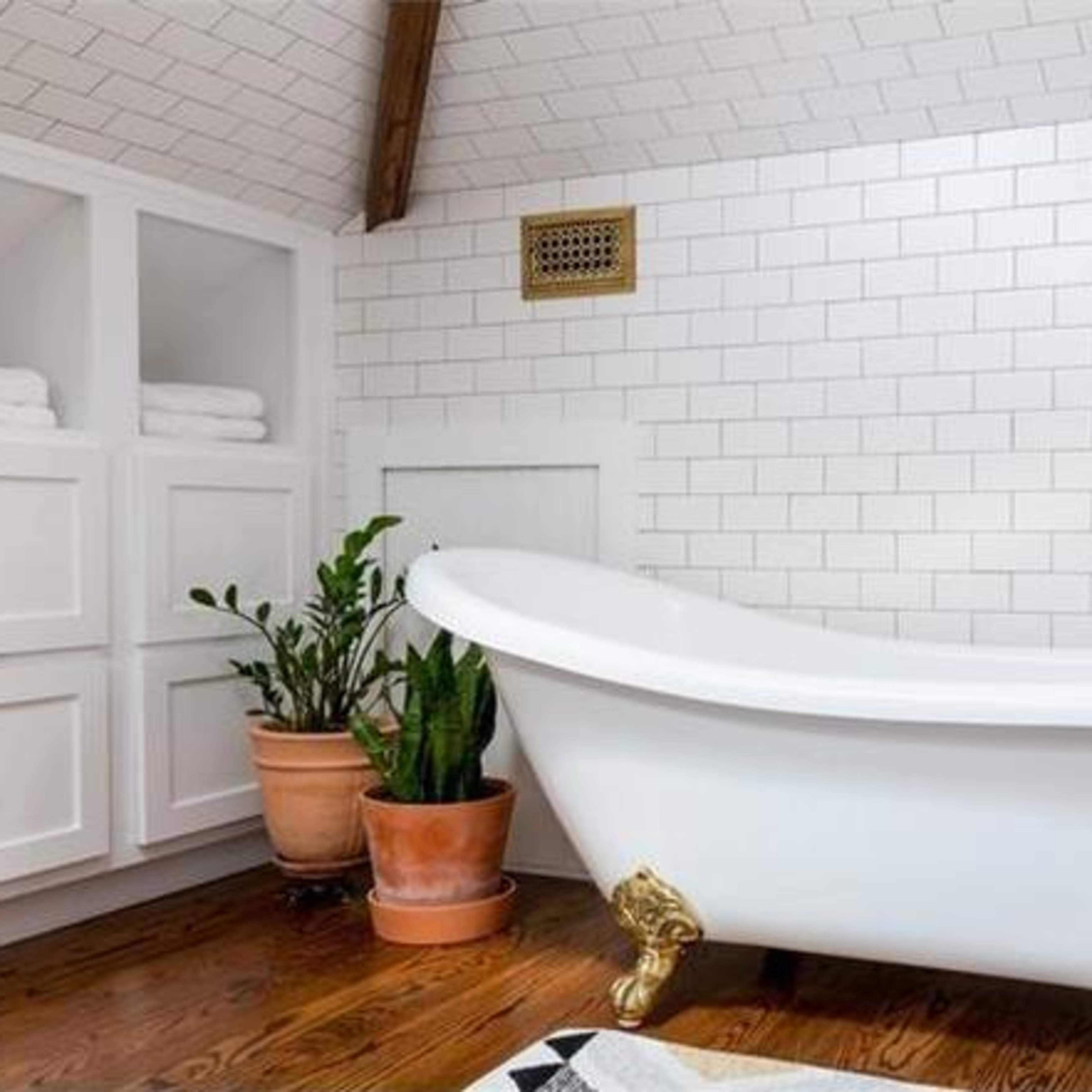 A bright bathroom featuring a white clawfoot bathtub, wooden flooring, and potted plants against a tiled wall.