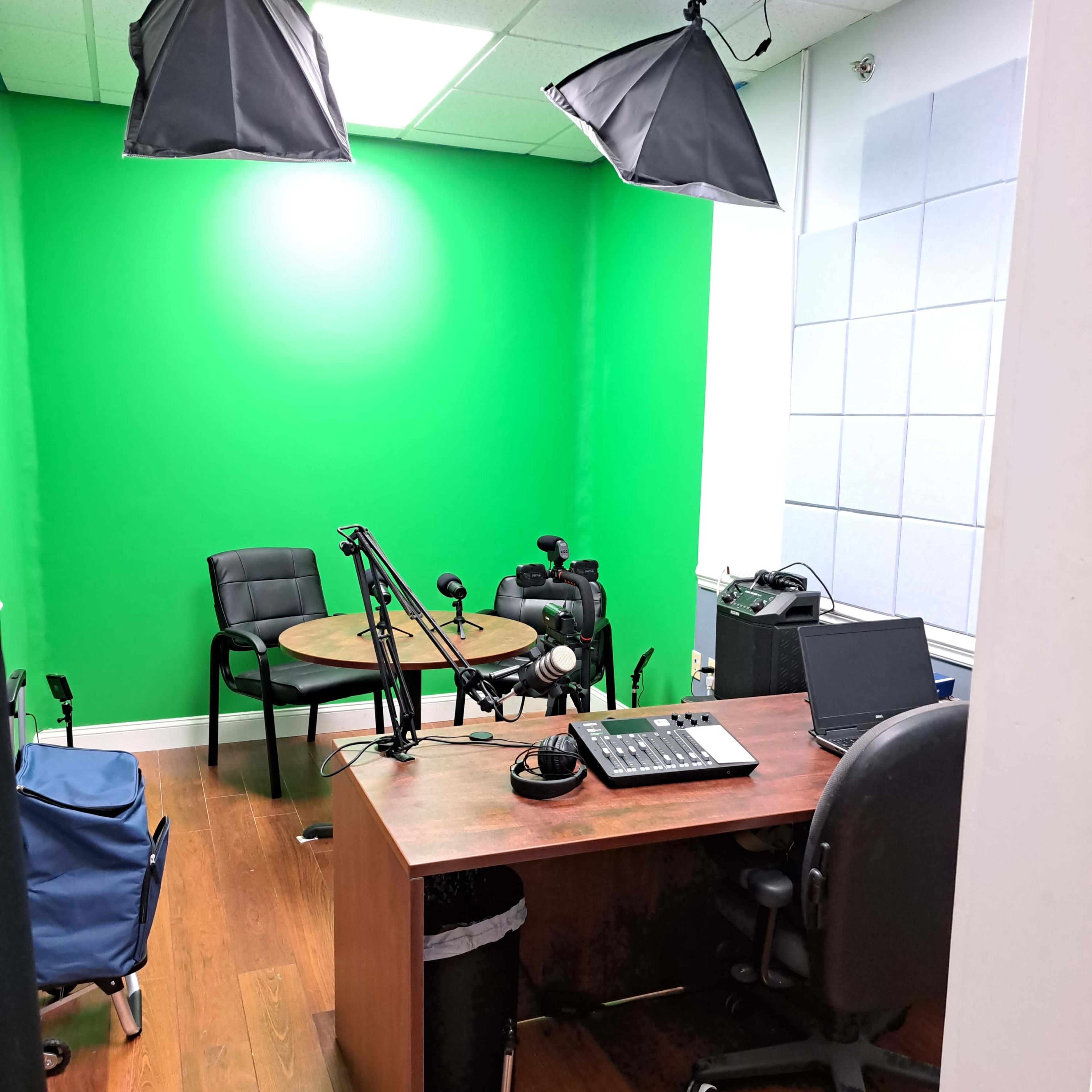 A recording studio with a green backdrop, a round table, chairs, microphones, and various recording equipment.