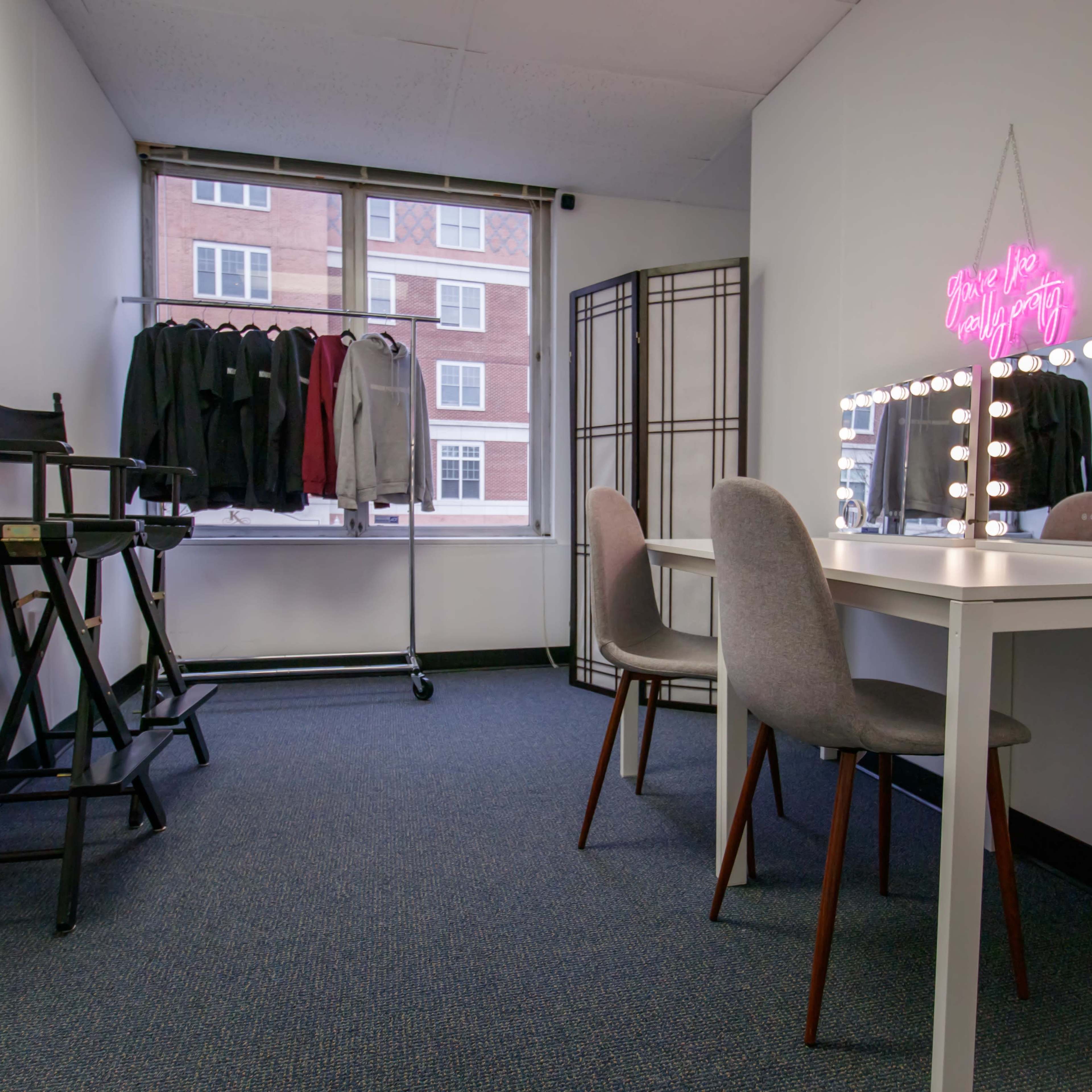 A well-lit room featuring a vanity mirror with lights, two gray chairs, a table, and a clothing rack with black garments near a window.