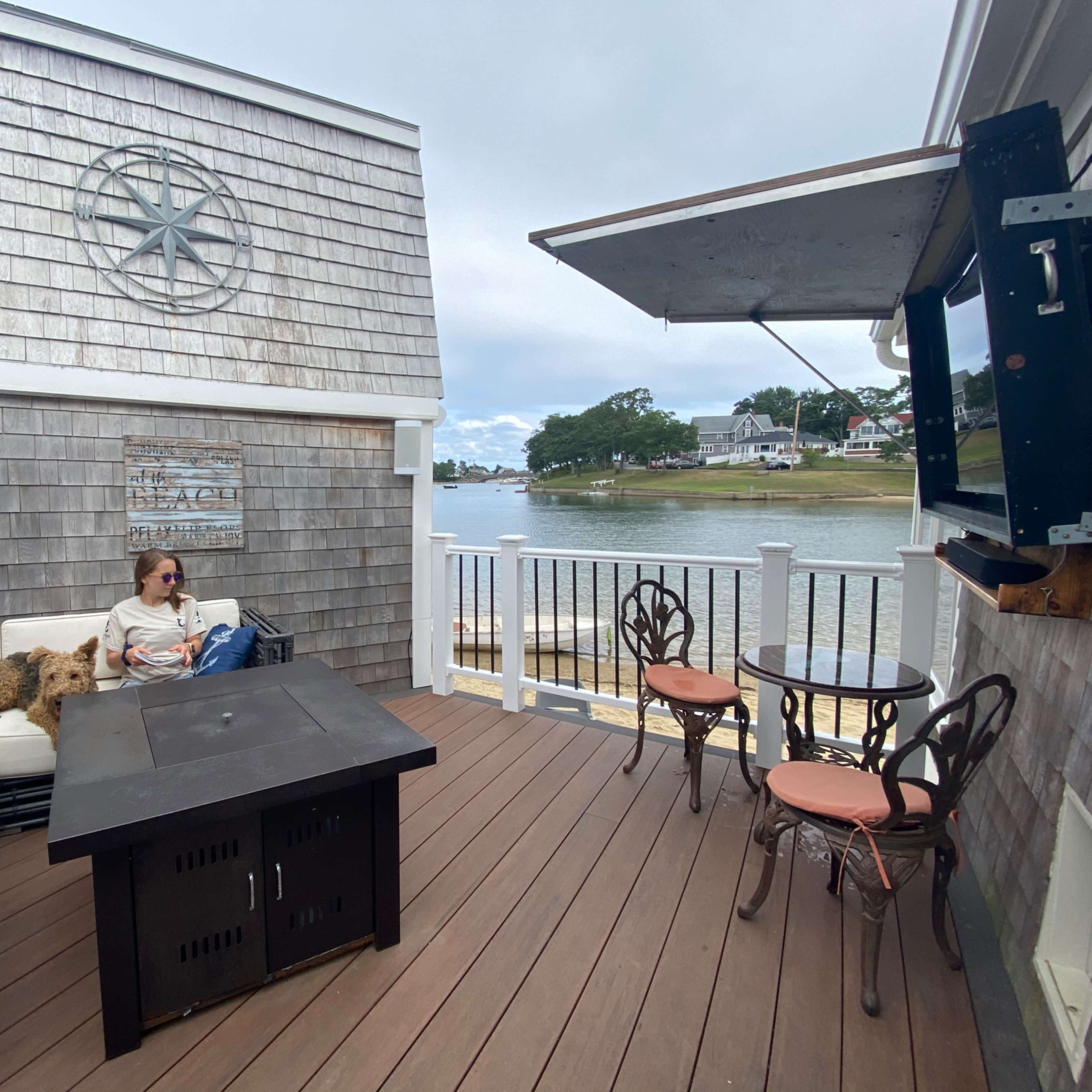 A person sits on a sofa on a deck overlooking a body of water, with a fire pit in the center and a TV mounted overhead.