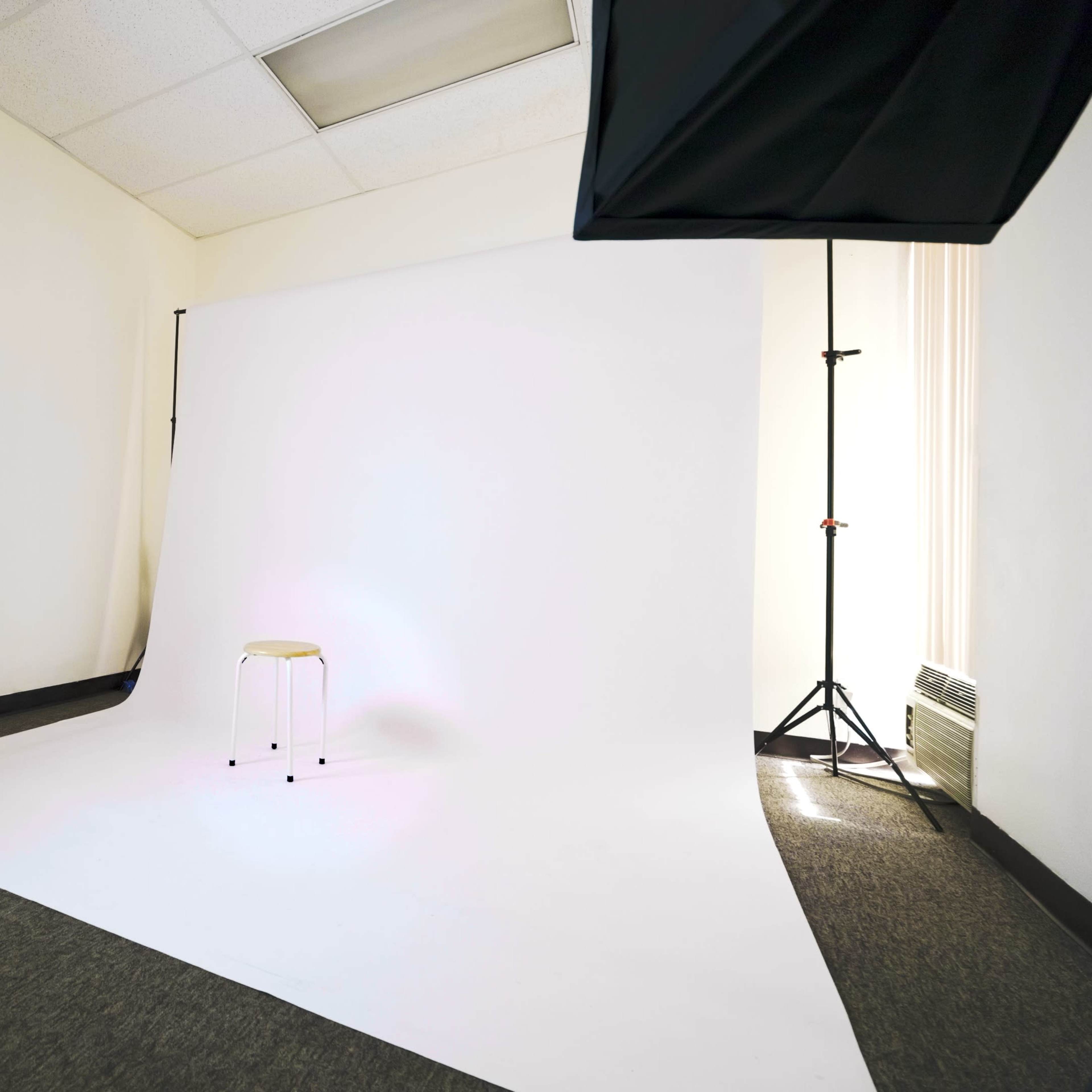 A photography studio with a white backdrop, a stool in the center, and professional lighting equipment set up.