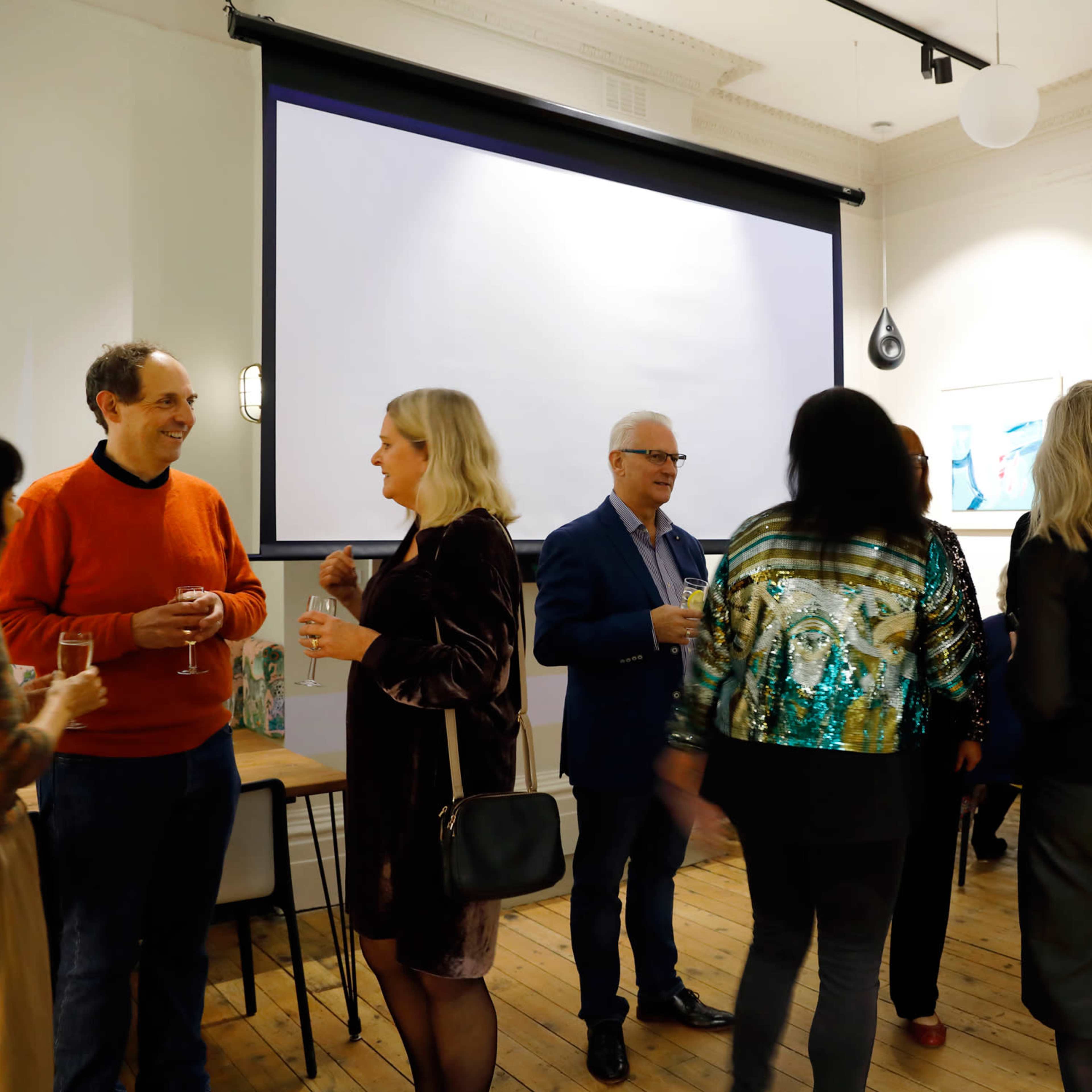 A group of people mingles in a well-lit social setting with a blank projector screen in the background.