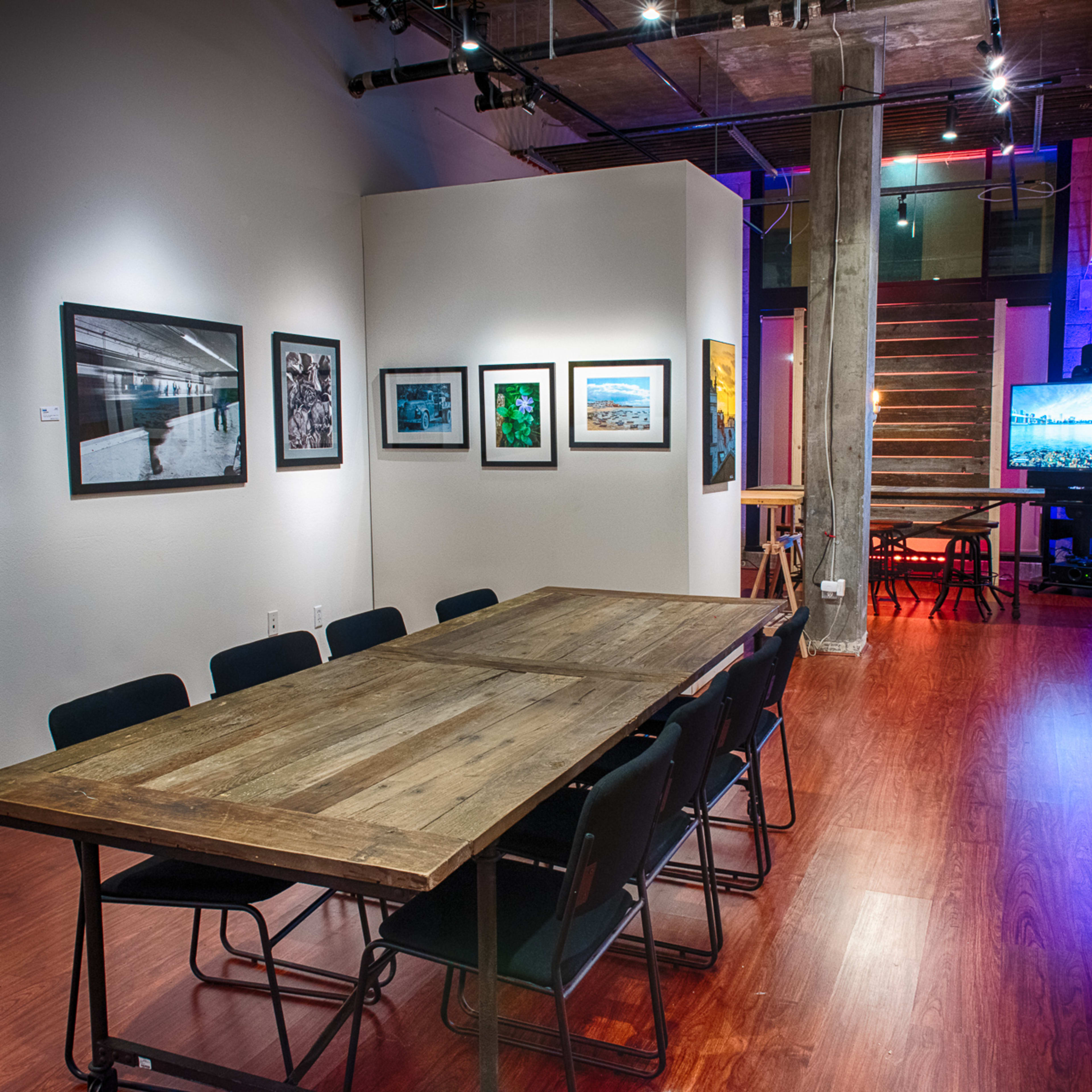 A spacious gallery features a long wooden table surrounded by black chairs, with framed photographs displayed on the walls and a staircase leading to an upper level.
