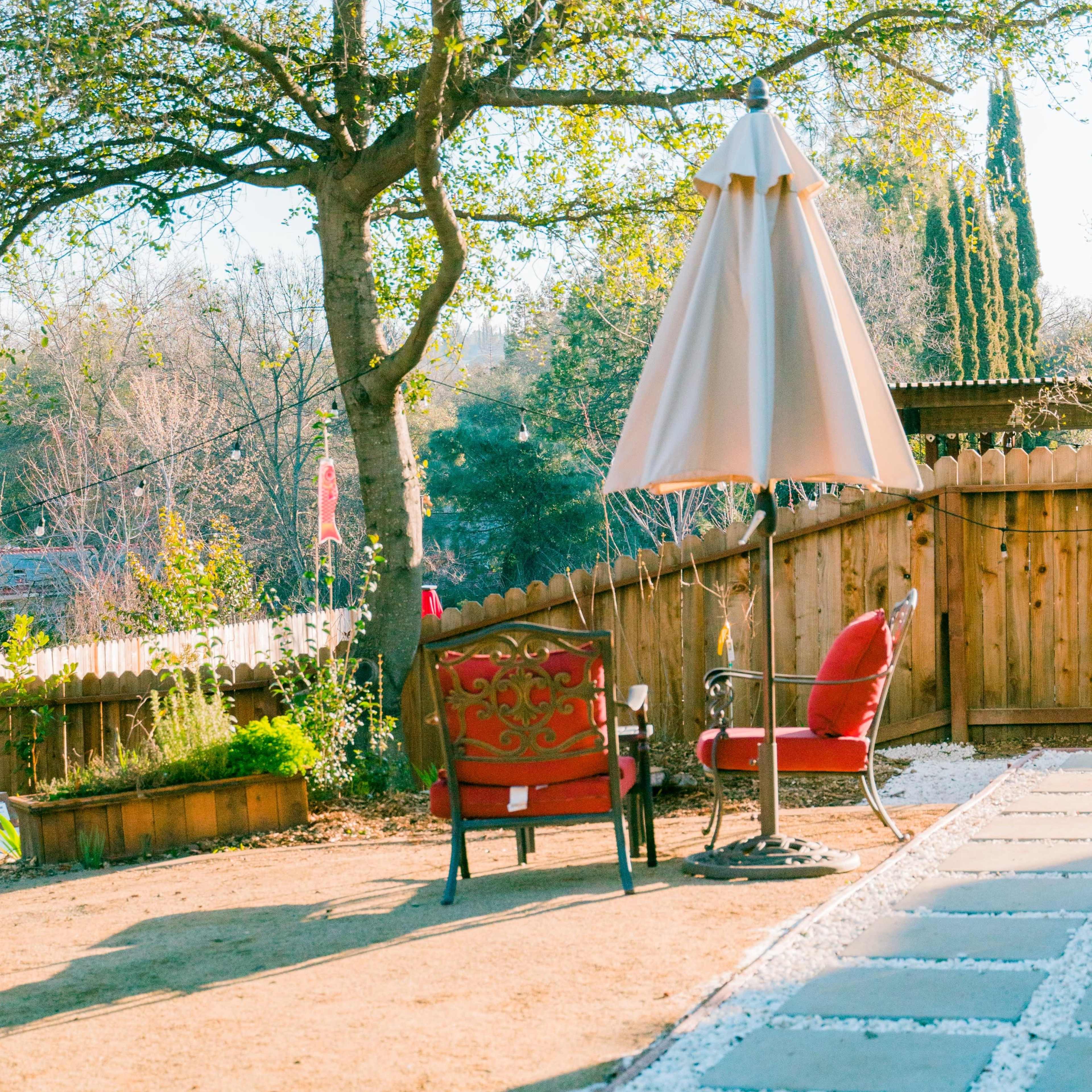 A patio area features two red chairs under a beige umbrella, surrounded by grass and a wooden fence.