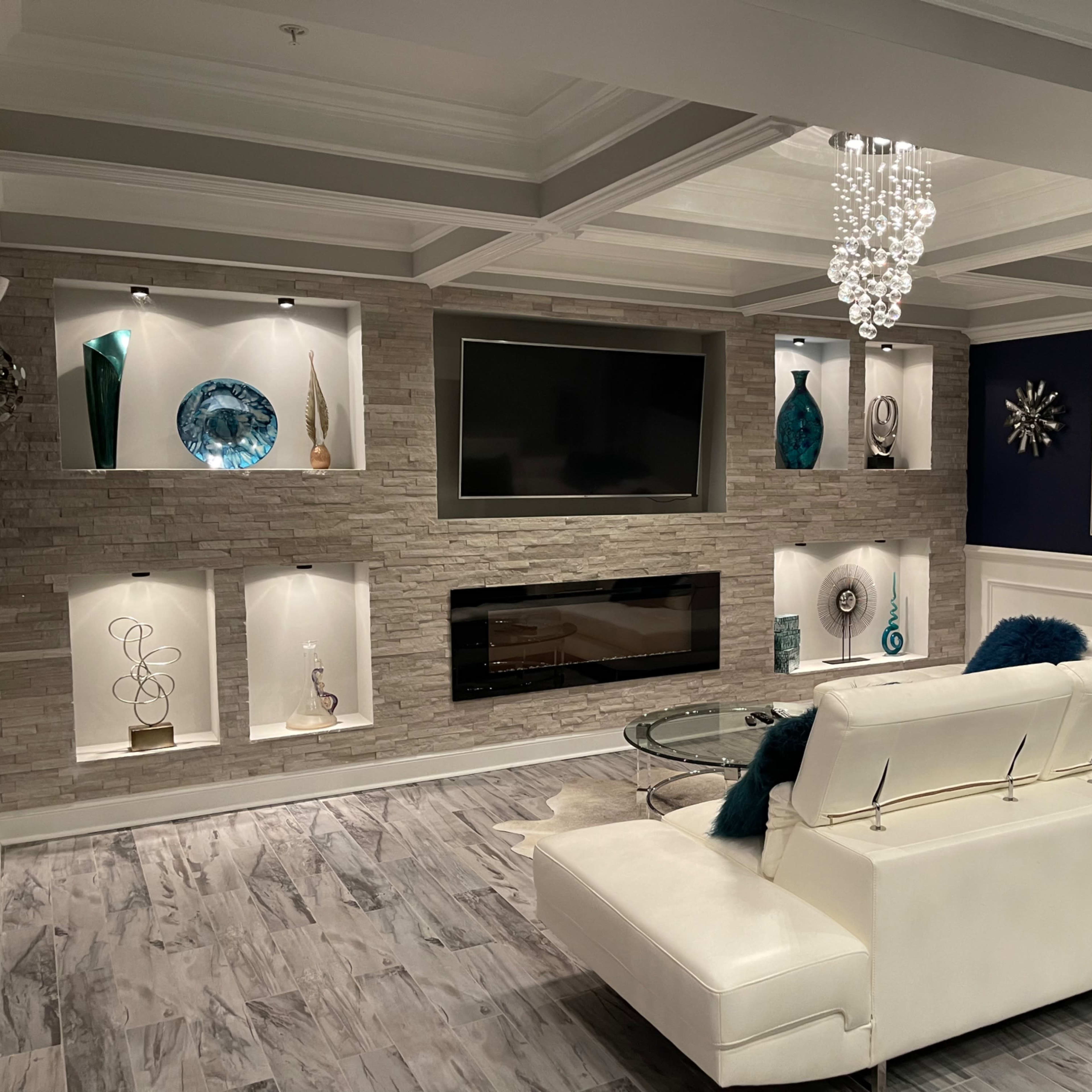 A modern living room featuring a white sectional sofa, a wall-mounted television, a stone accent wall with decorative shelves, and a glass coffee table under a chandelier.