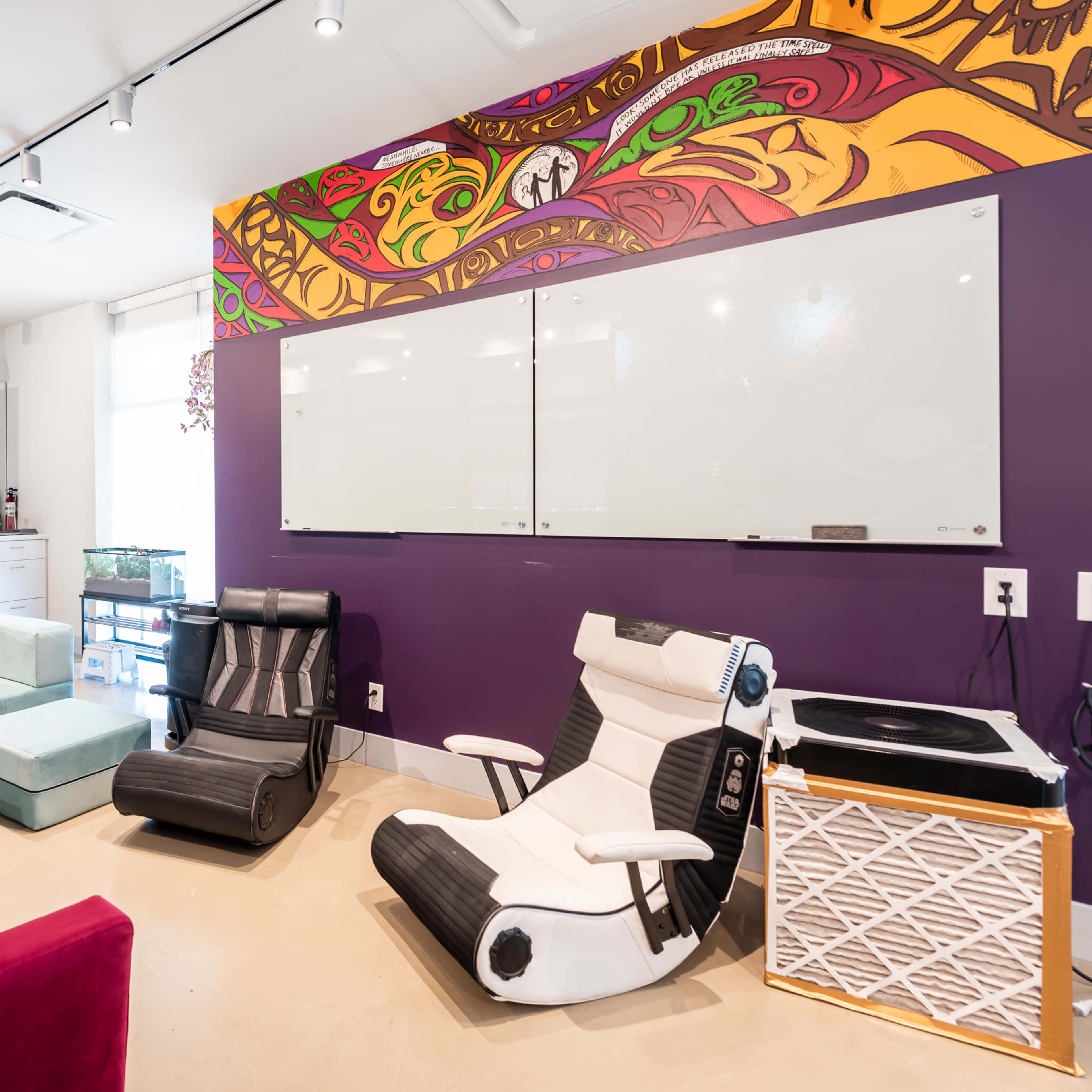 A modern lounge area with colorful walls, seating options including a red velvet couch and two lounge chairs, and a kitchenette in the background.