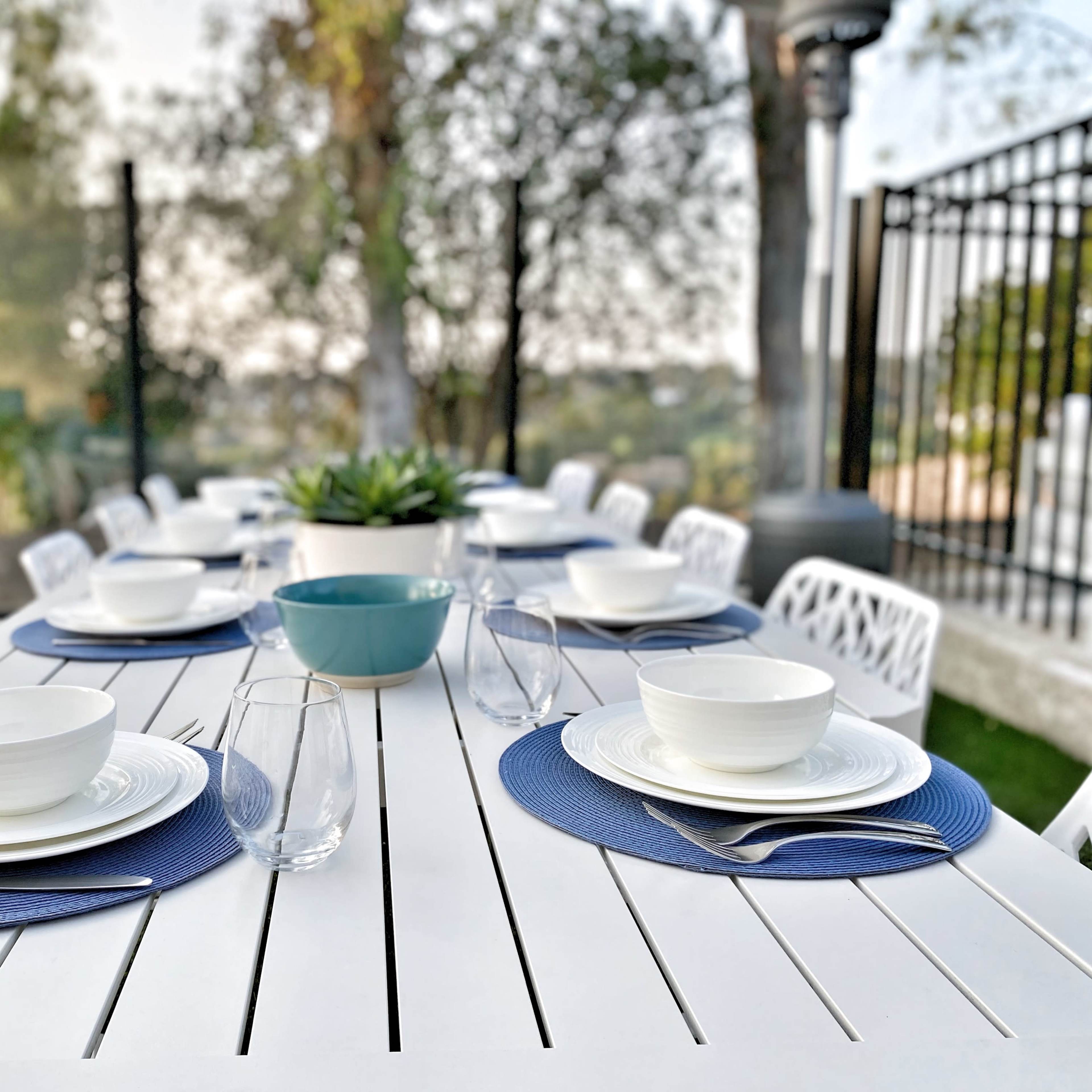 A long outdoor dining table is set with white plates, blue bowls, glassware, and navy placemats, surrounded by white chairs in a garden setting.