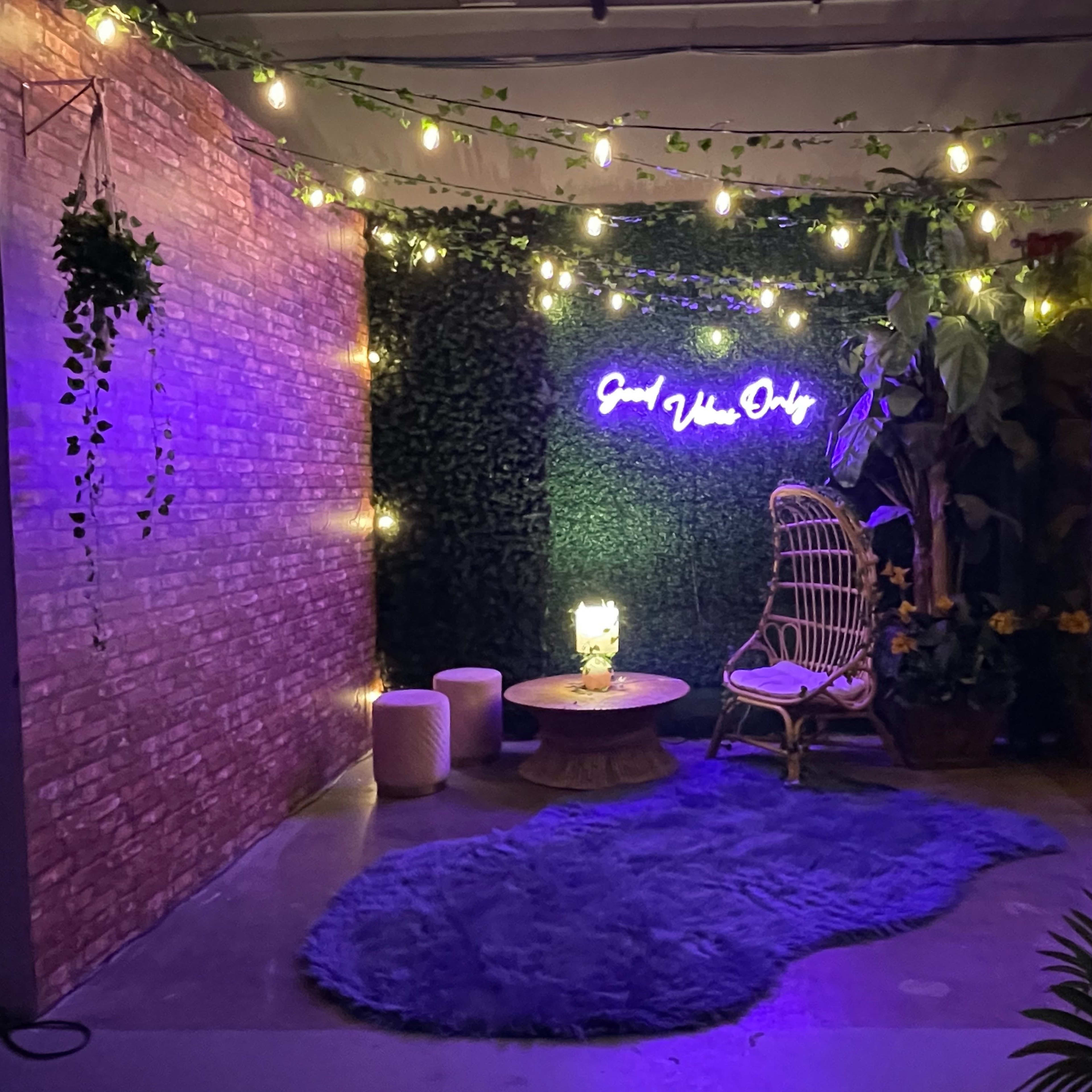 A cozy indoor space featuring a rattan chair, a round table, a blue rug, and string lights, with a green wall and a neon sign that reads "Good Vibes Only."