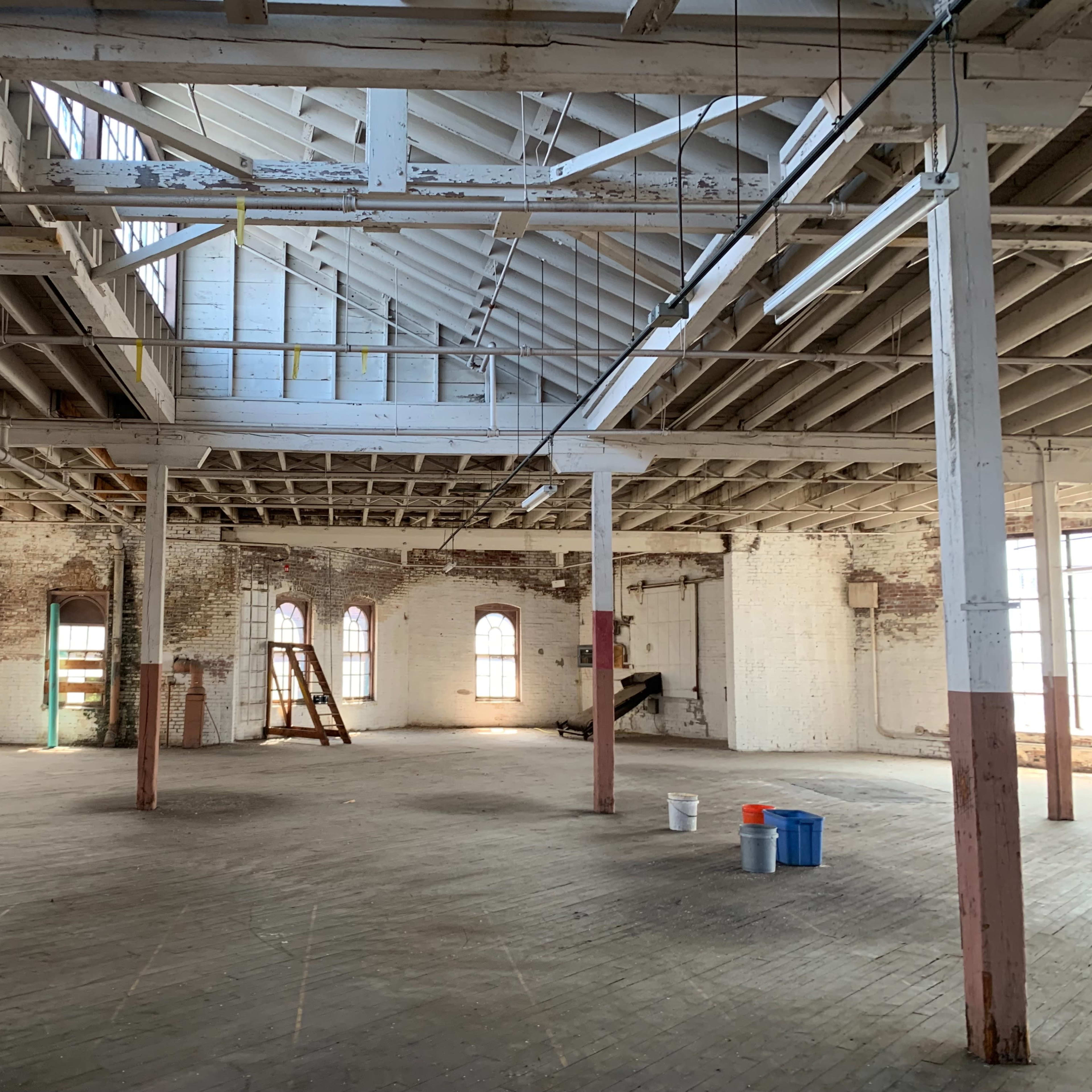 An empty industrial space with high ceilings, exposed beams, and several large windows.