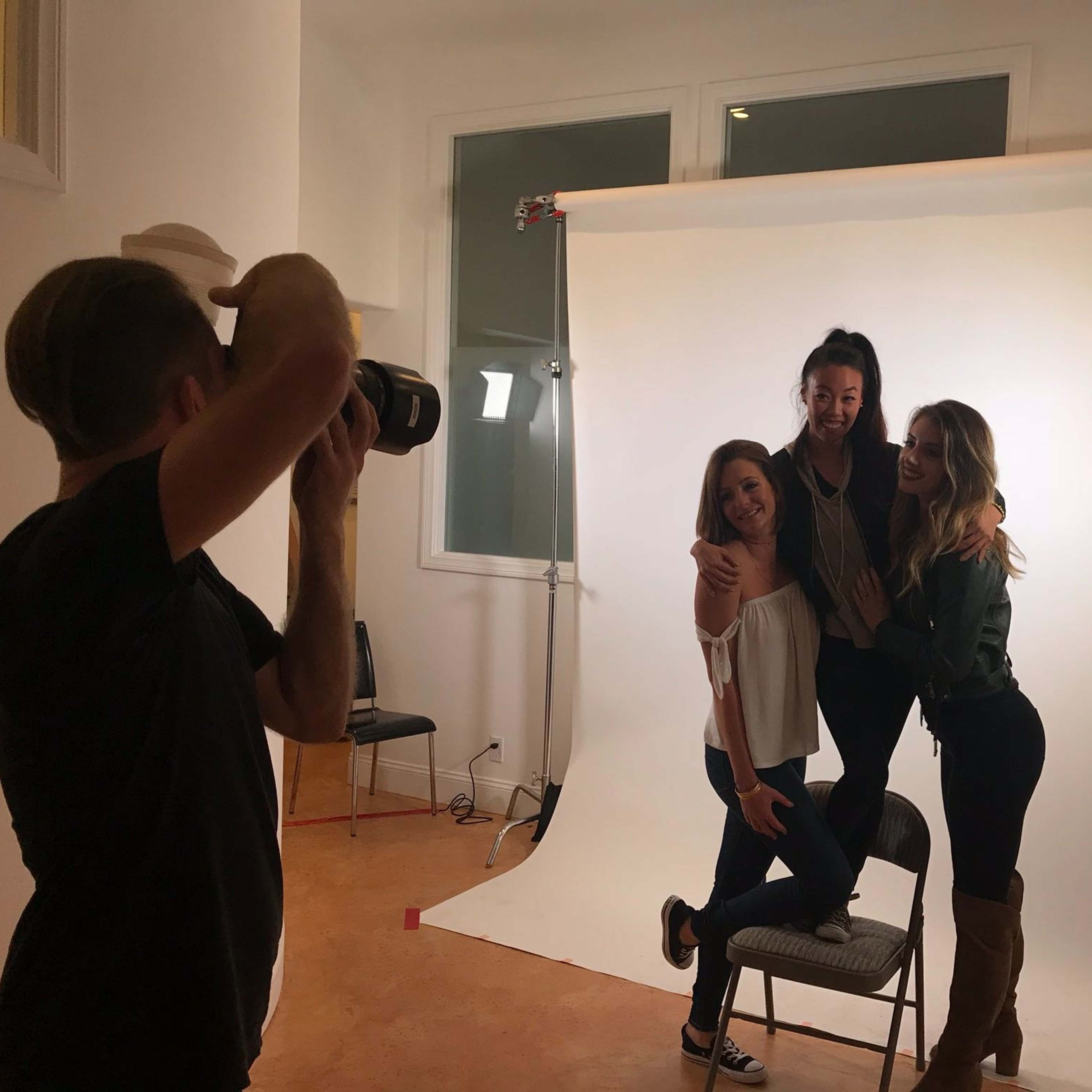 Three women pose together on a set with a white backdrop while a photographer takes their picture.