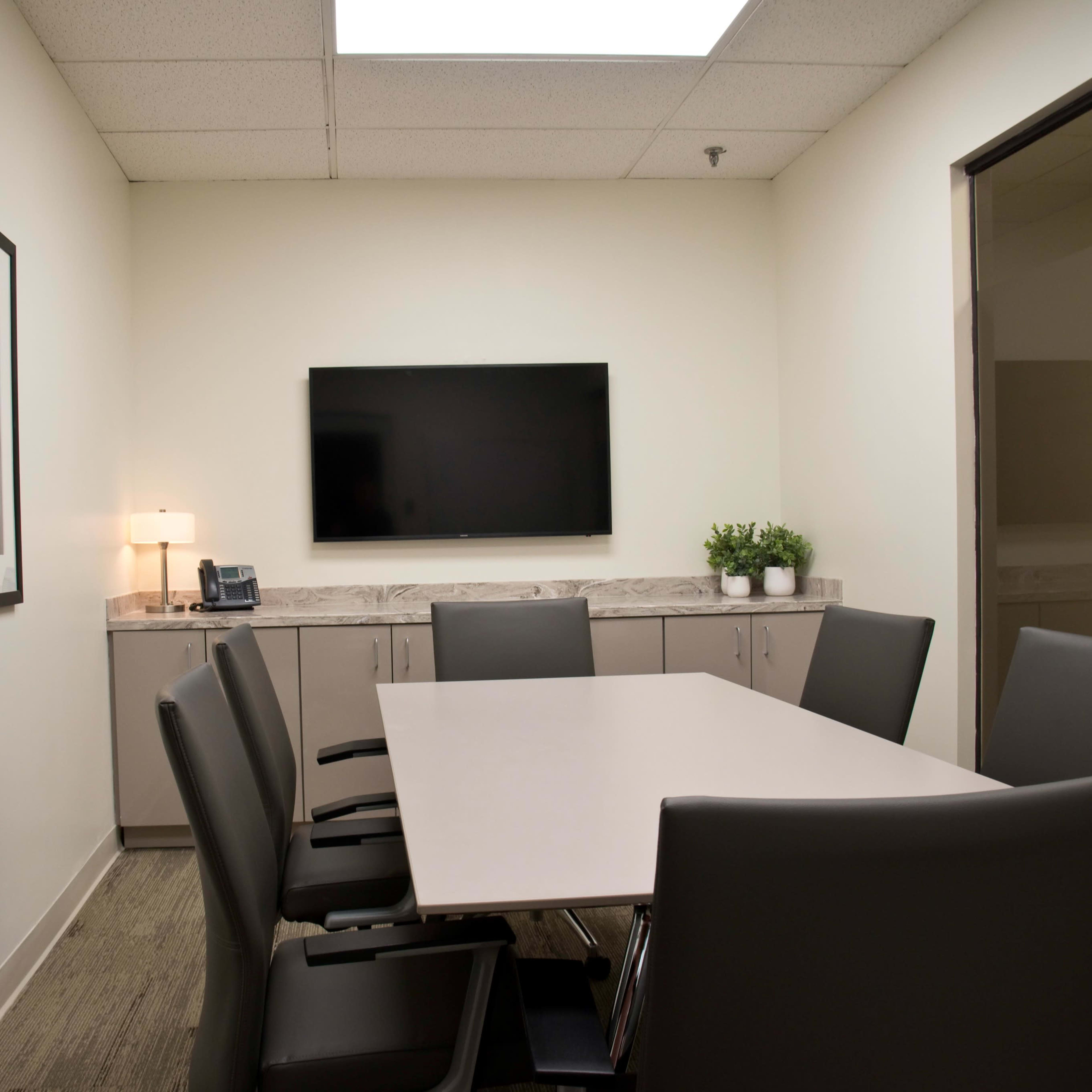 A modern conference room with a rectangular table surrounded by six chairs, a large wall-mounted television, and a cabinet with a lamp and plants.