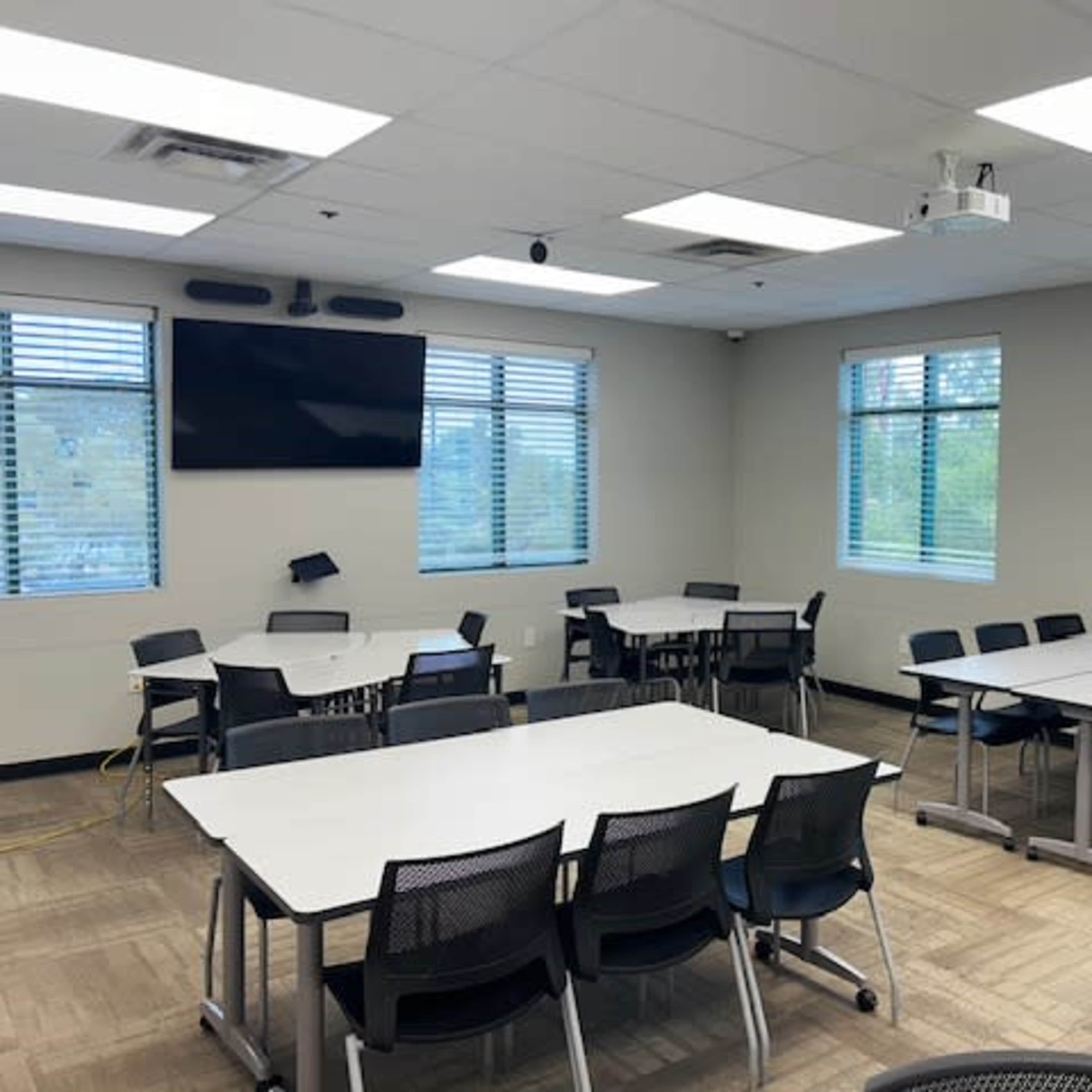 A well-lit classroom features multiple tables and chairs arranged for meetings or group work, with a projector and screen mounted on the wall.