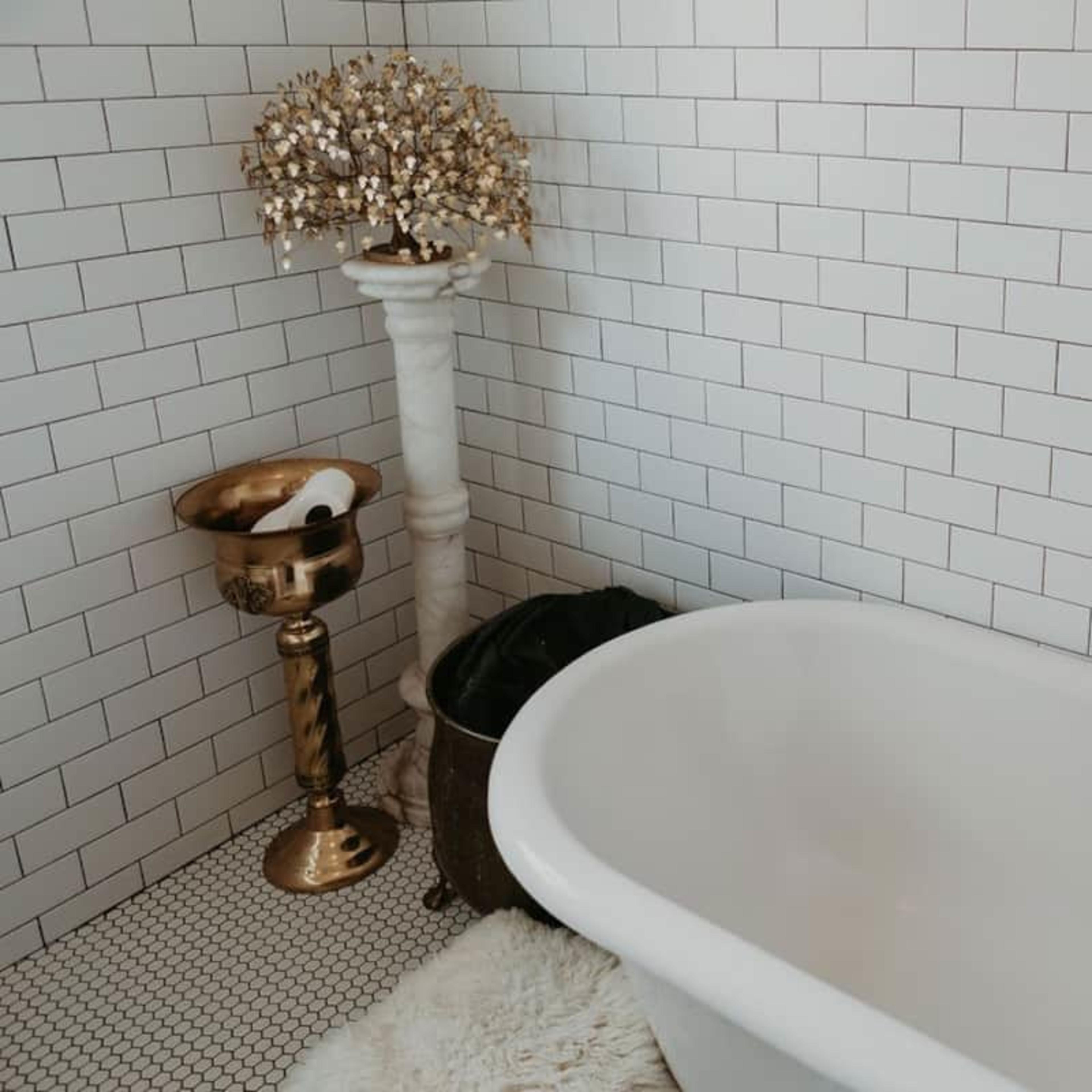 A white bathtub with a wooden bath tray beside a decorative pedestal and a gold bowl holding rolled towels in a tiled bathroom.