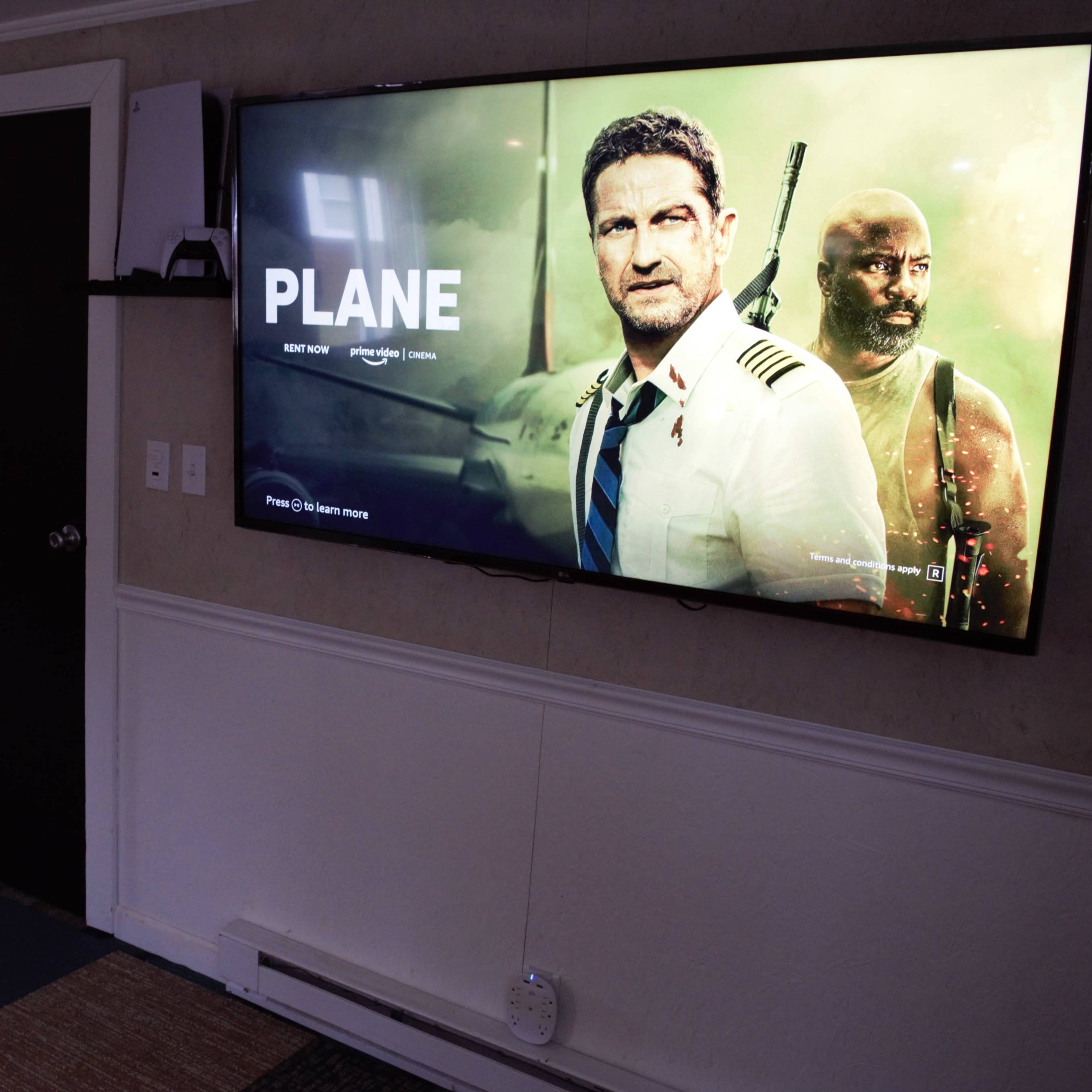 A large television displays the movie "Plane," featuring a male actor in a pilot uniform alongside another character in a military uniform, set in a living room with dark doors and a carpeted floor.