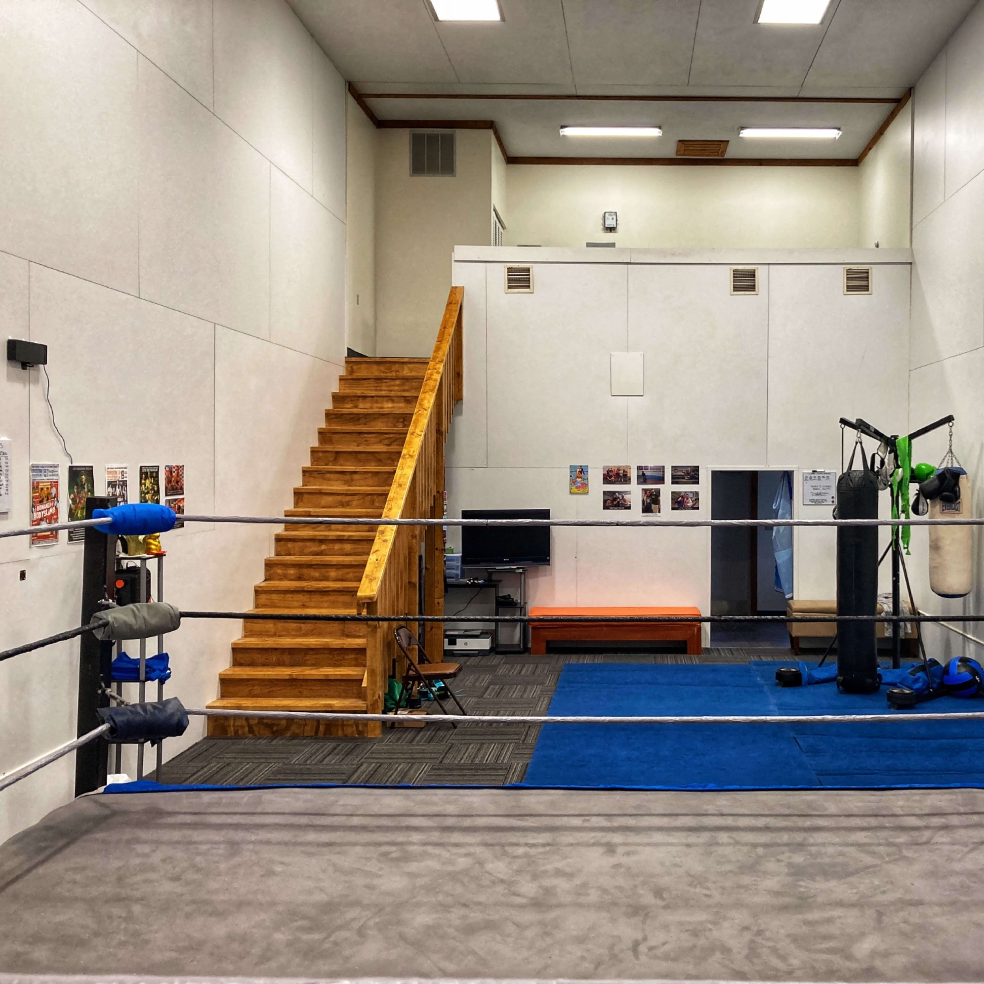 A training facility with a boxing ring in the foreground, a set of stairs on the left, and various workout equipment and posters on the walls.