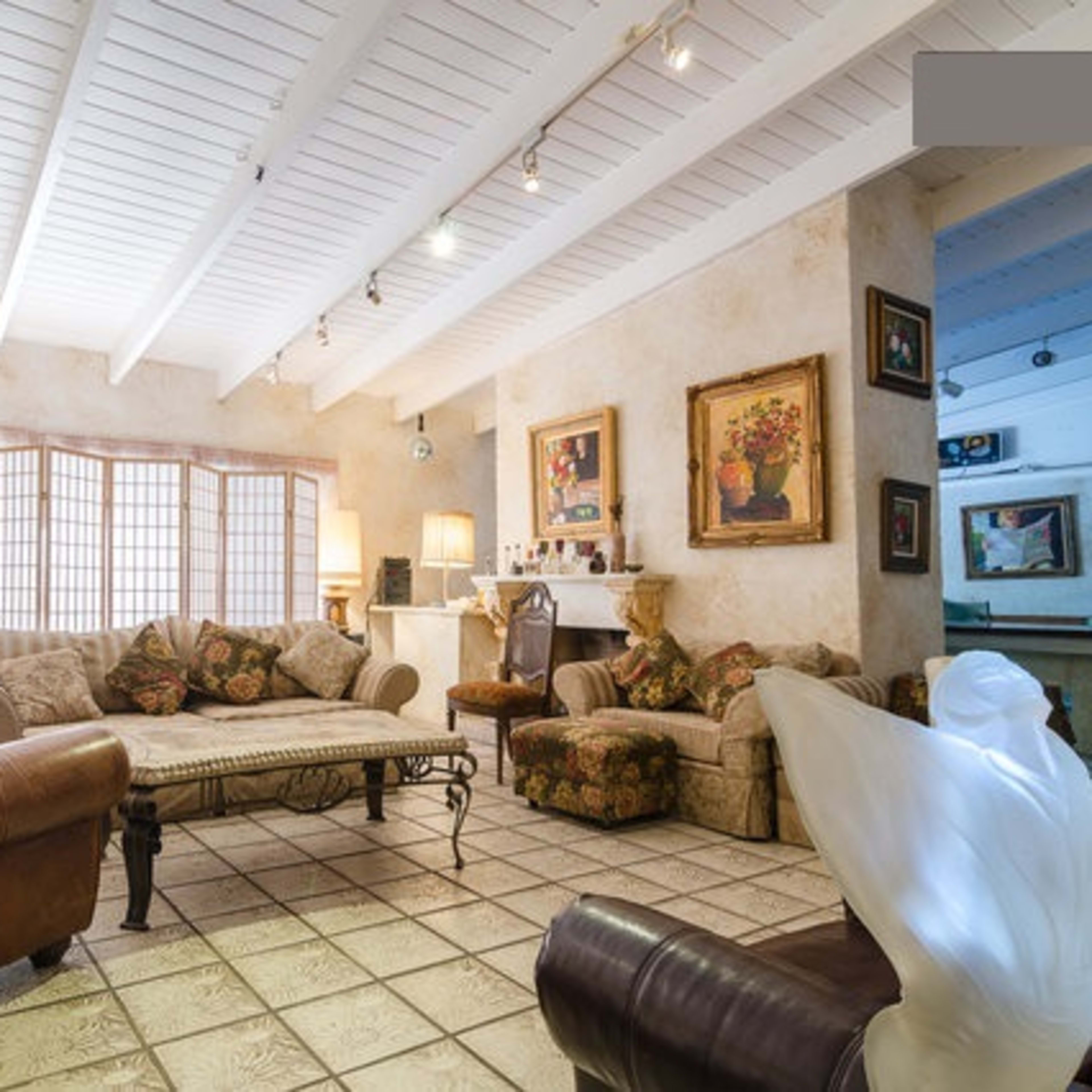 A spacious living room with a patterned sofa, several artworks on the walls, and a large window allowing natural light to enter.