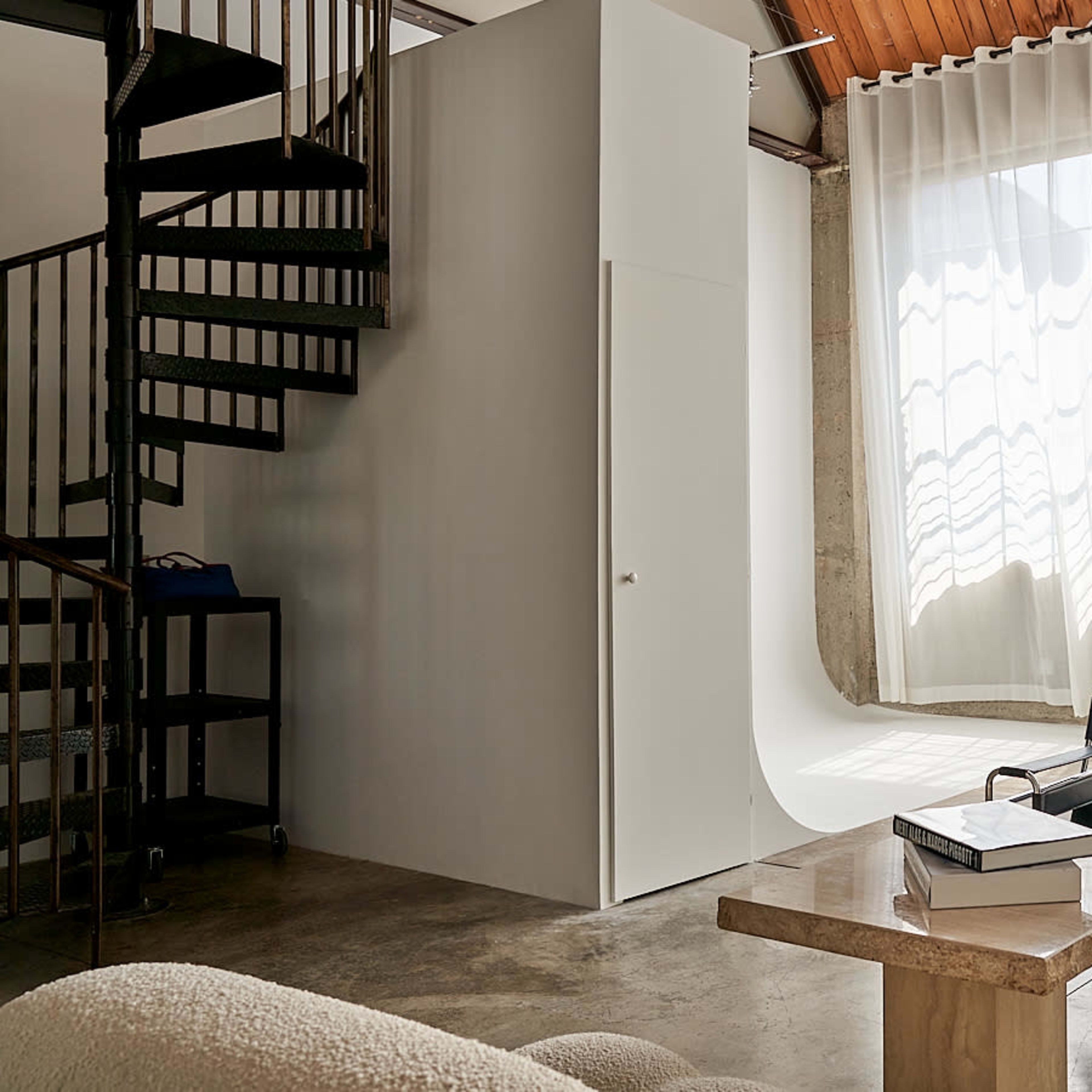 A modern interior space with a spiral staircase, large windows featuring sheer curtains, and minimalist furniture arranged around a central table.