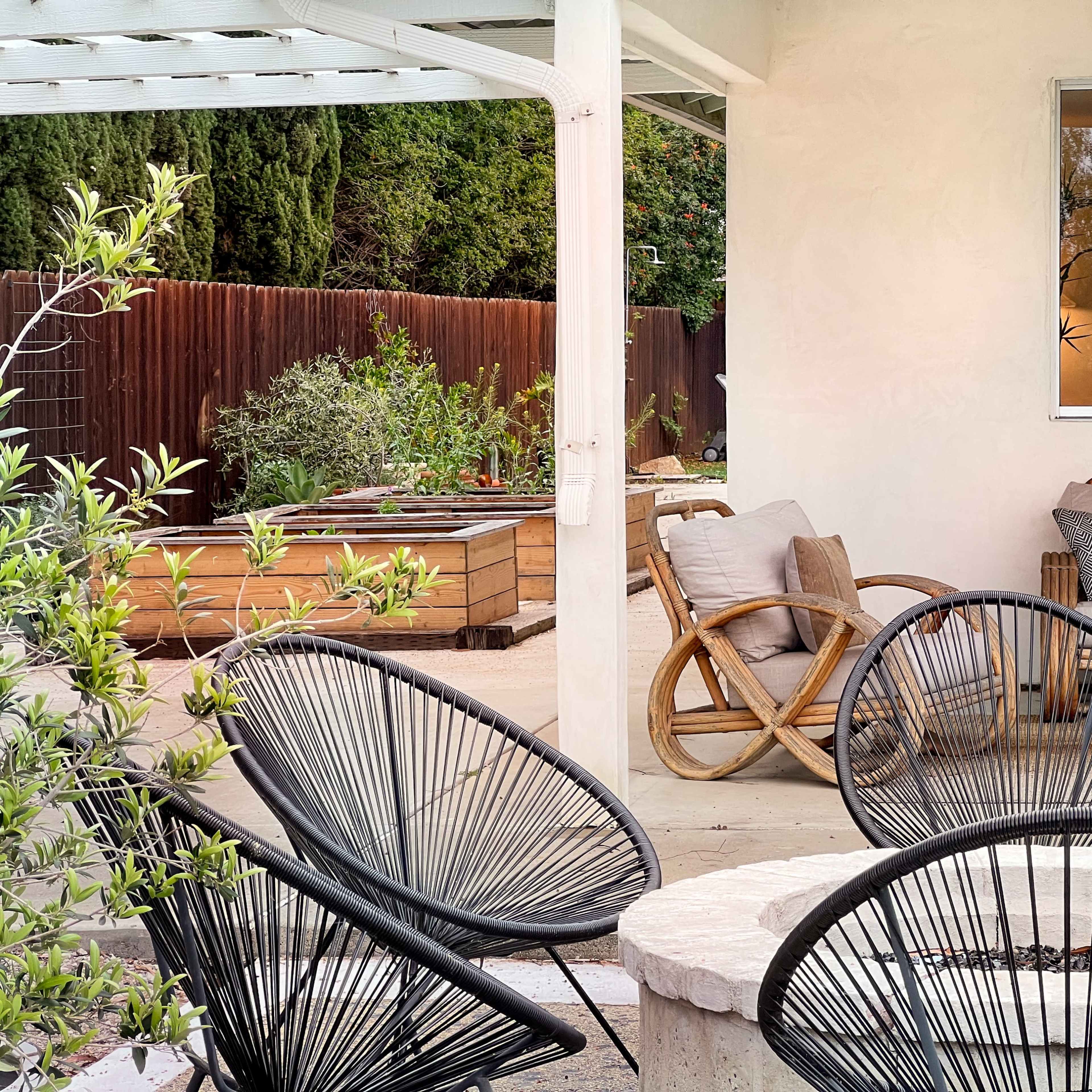 A cozy outdoor patio area with various seating arrangements, including black wire chairs and a wooden couch, surrounded by plants and a fire pit.