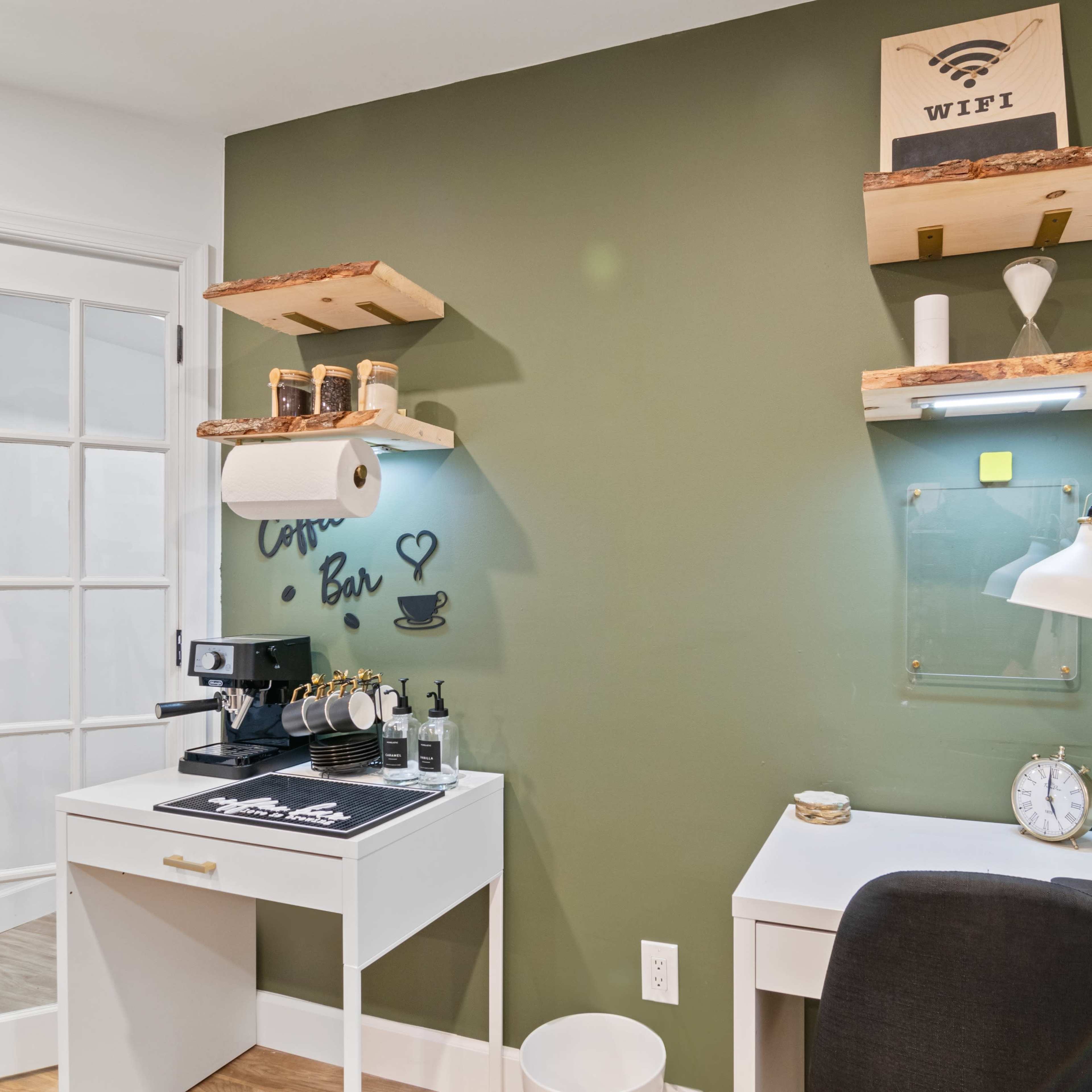 A small workspace with a coffee machine on a white desk, shelves mounted on a green wall, and a bright desk lamp.