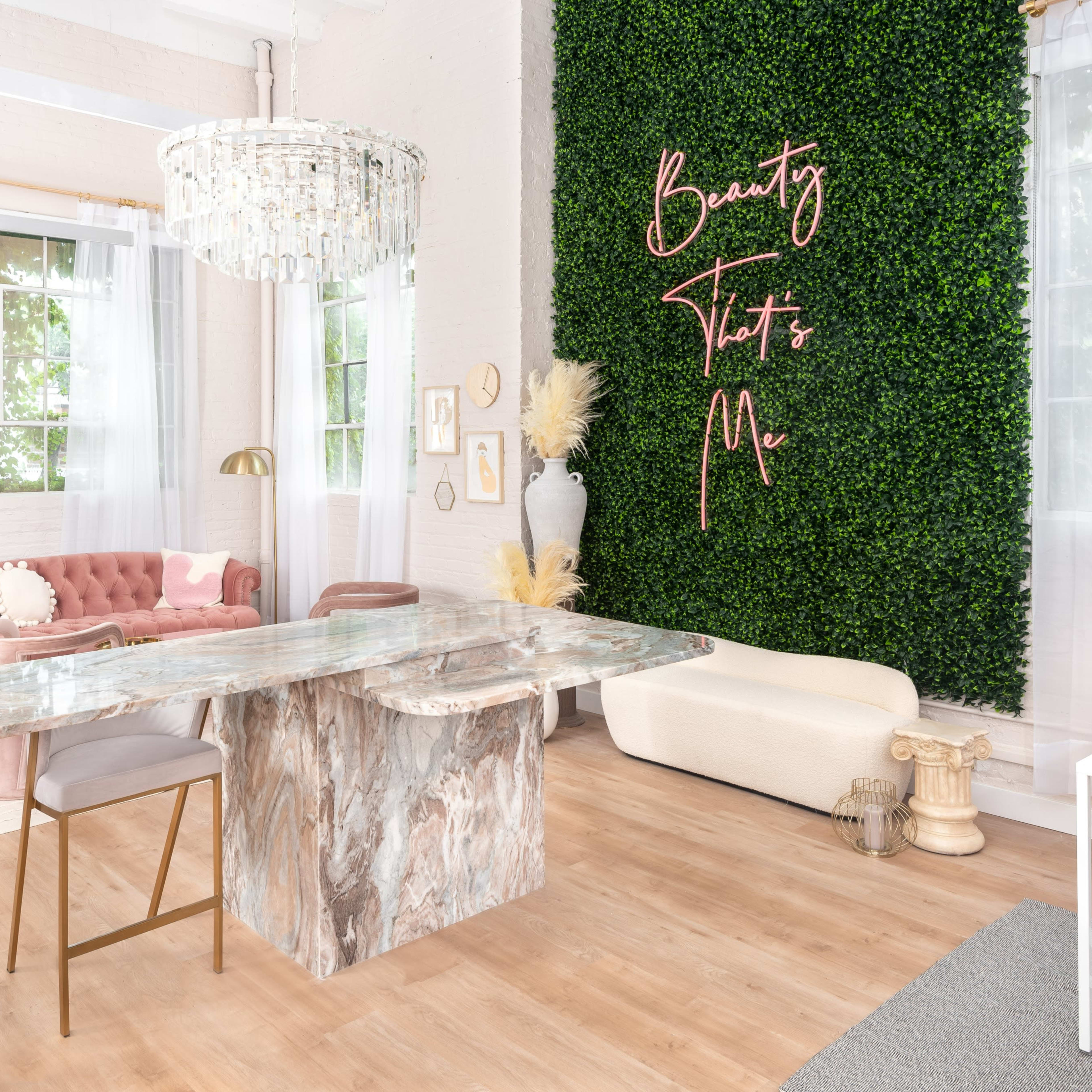 A modern interior space featuring a marble table, a pink velvet sofa, and a greenery wall with the words "Beauty That's Me" in elegant lettering.