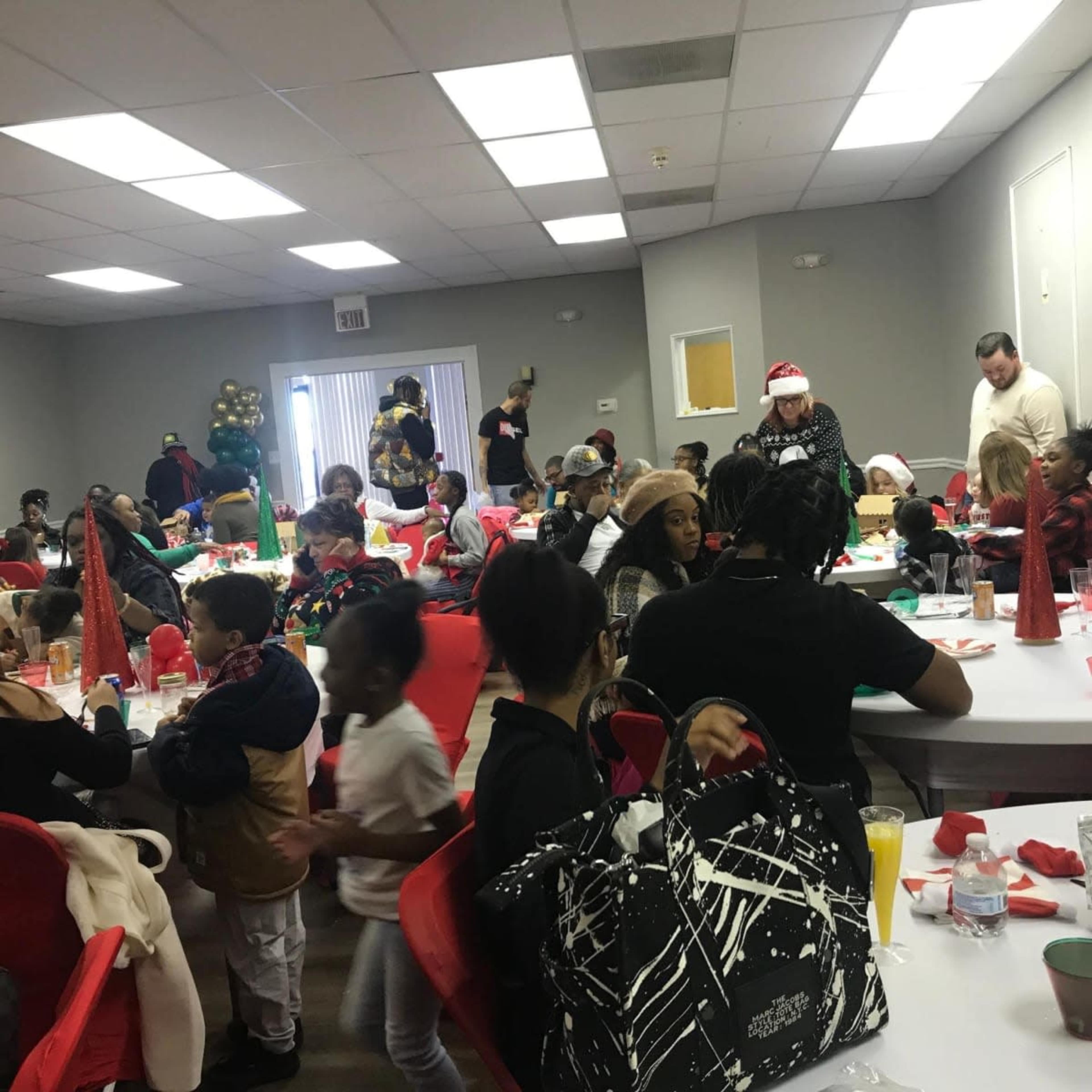 A festive gathering is taking place in a large room filled with tables, people chatting, and holiday decorations.