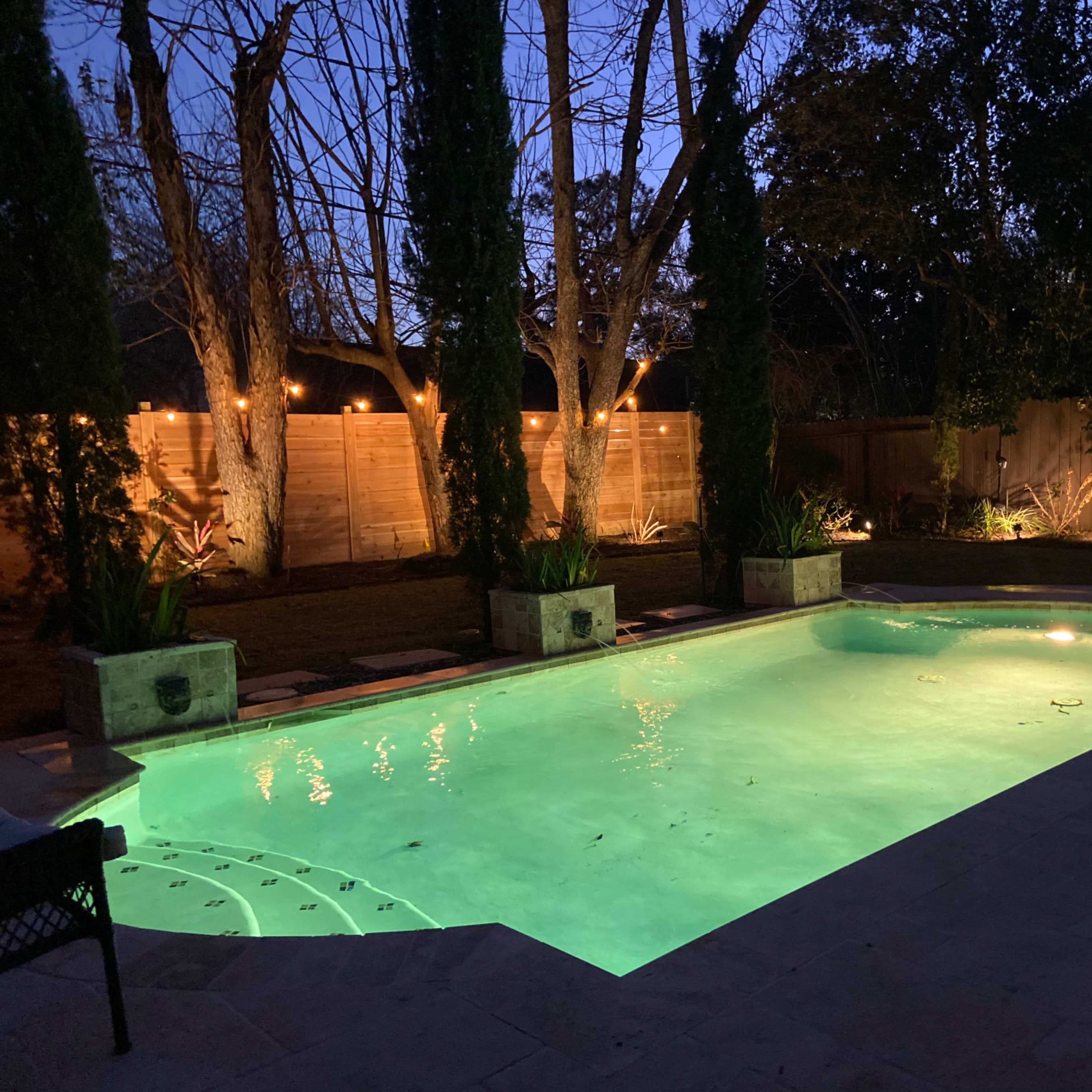 A well-lit swimming pool surrounded by landscaped trees and a wooden fence, with lights illuminating the area at dusk.