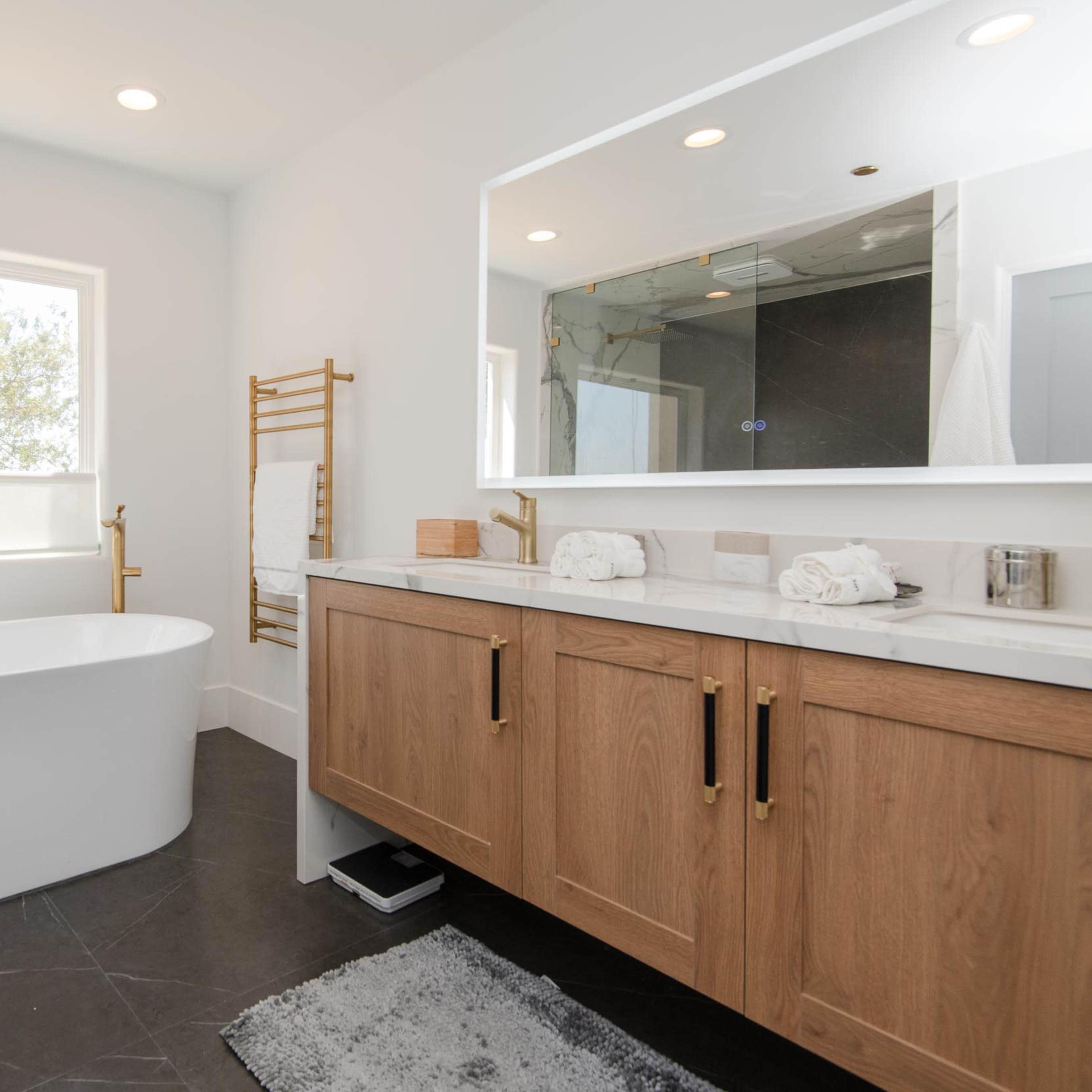 The bathroom features a freestanding white bathtub, a wooden vanity with brushed gold fixtures, and a large mirror above it, along with a window letting in natural light.