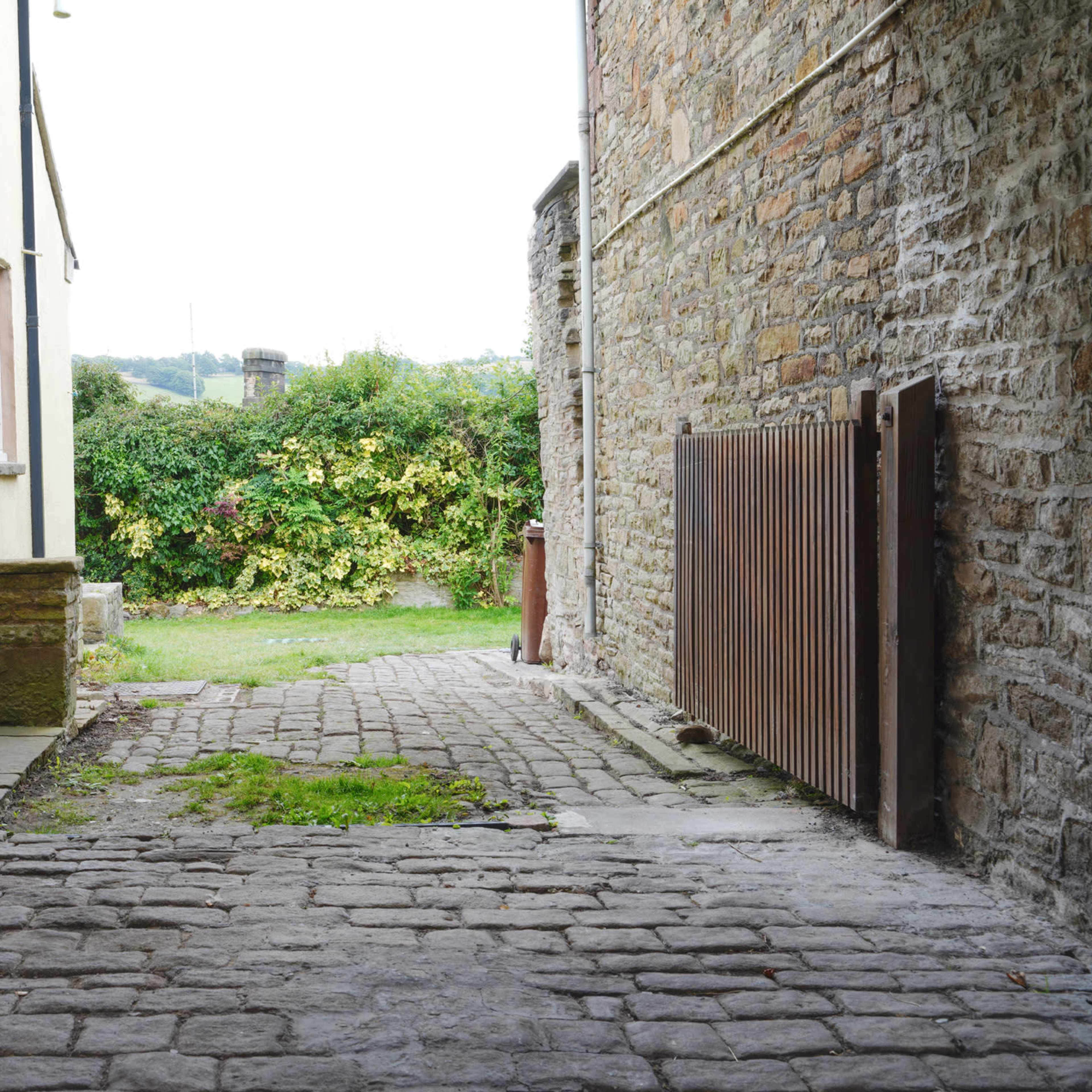 A cobbled pathway between two stone buildings, leading to a green garden visible in the background.