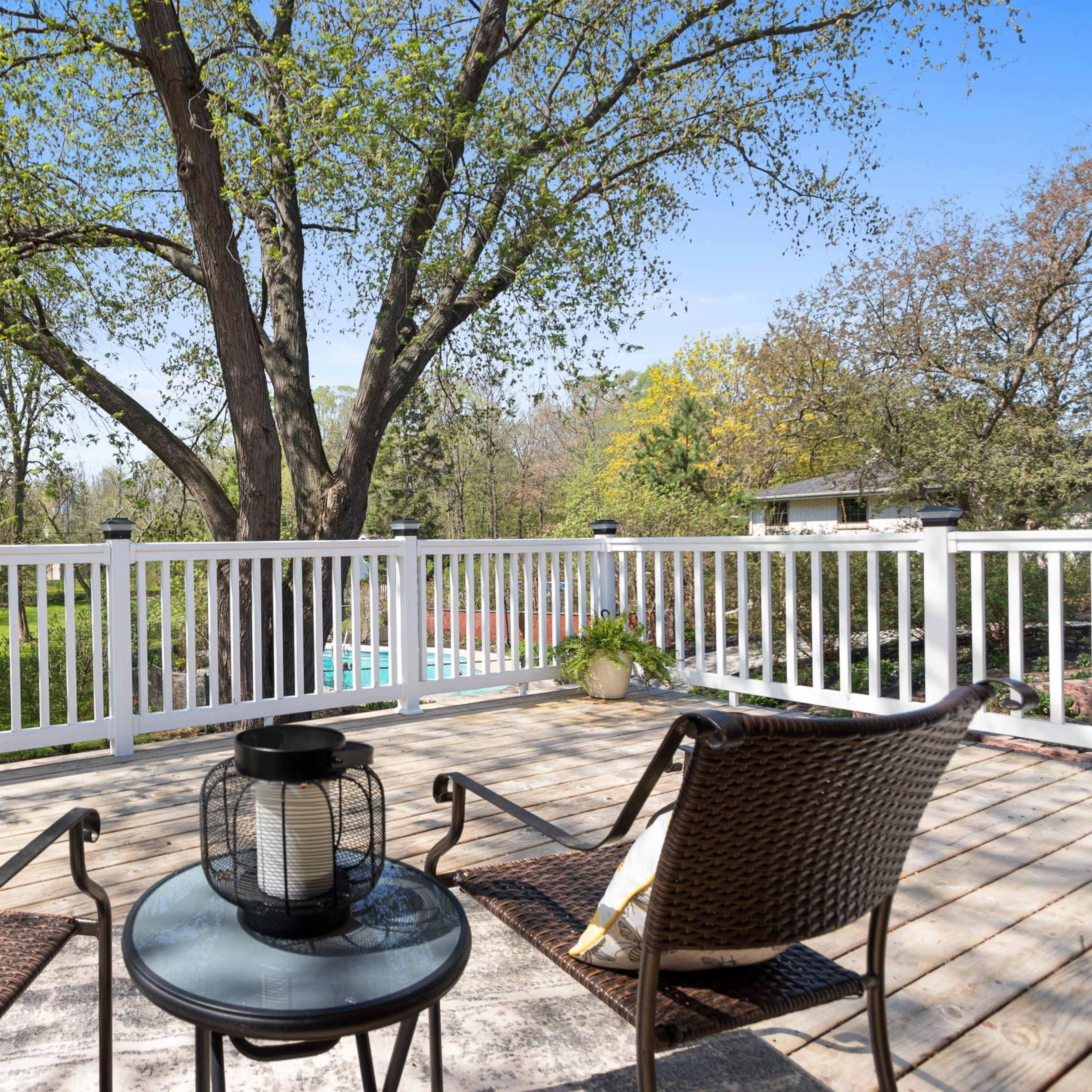 A deck with two wicker chairs and a black lantern overlooks a green yard with trees and a swimming pool in the background.