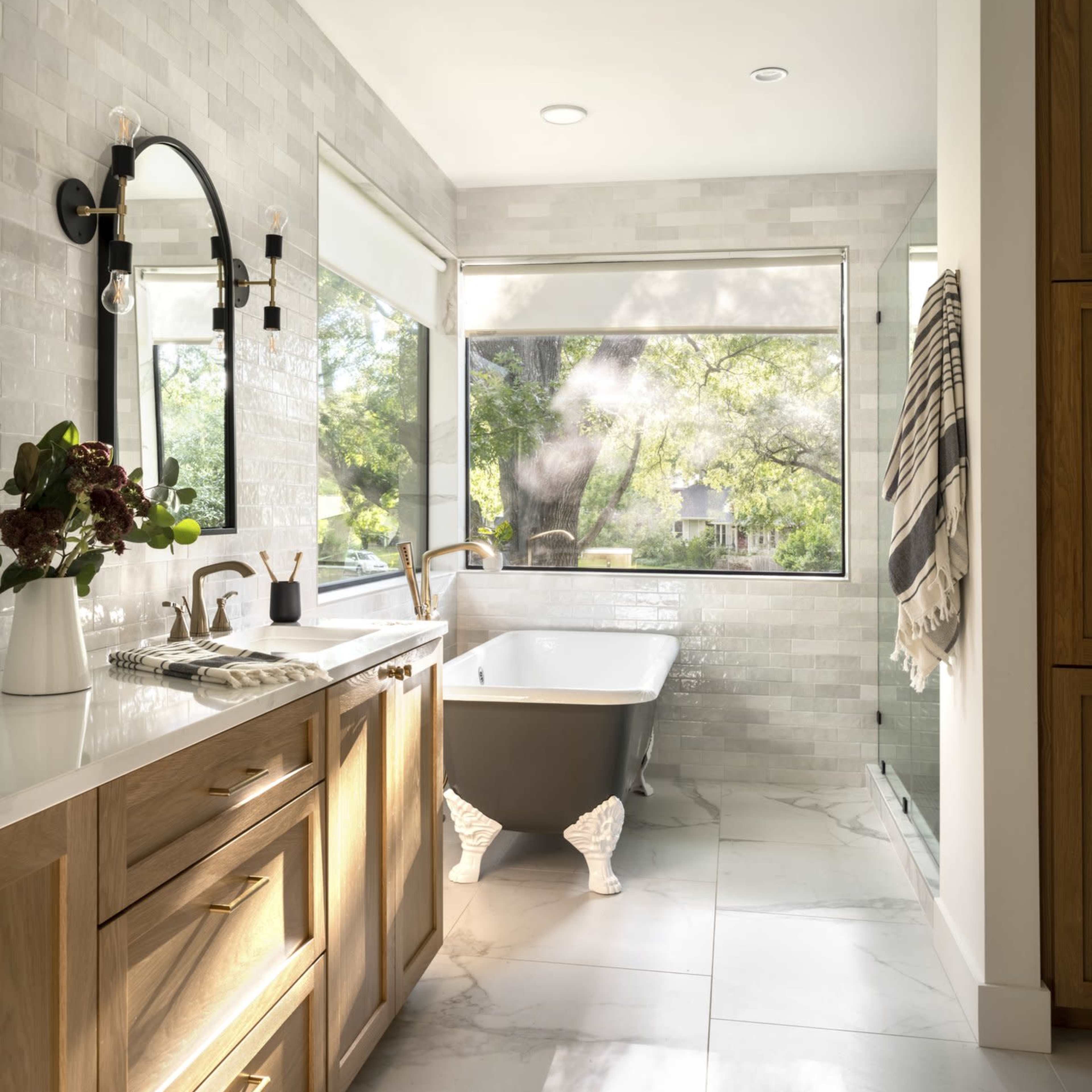 A modern bathroom features a freestanding clawfoot tub, large window with a view, wooden cabinetry, and a glass shower enclosure.