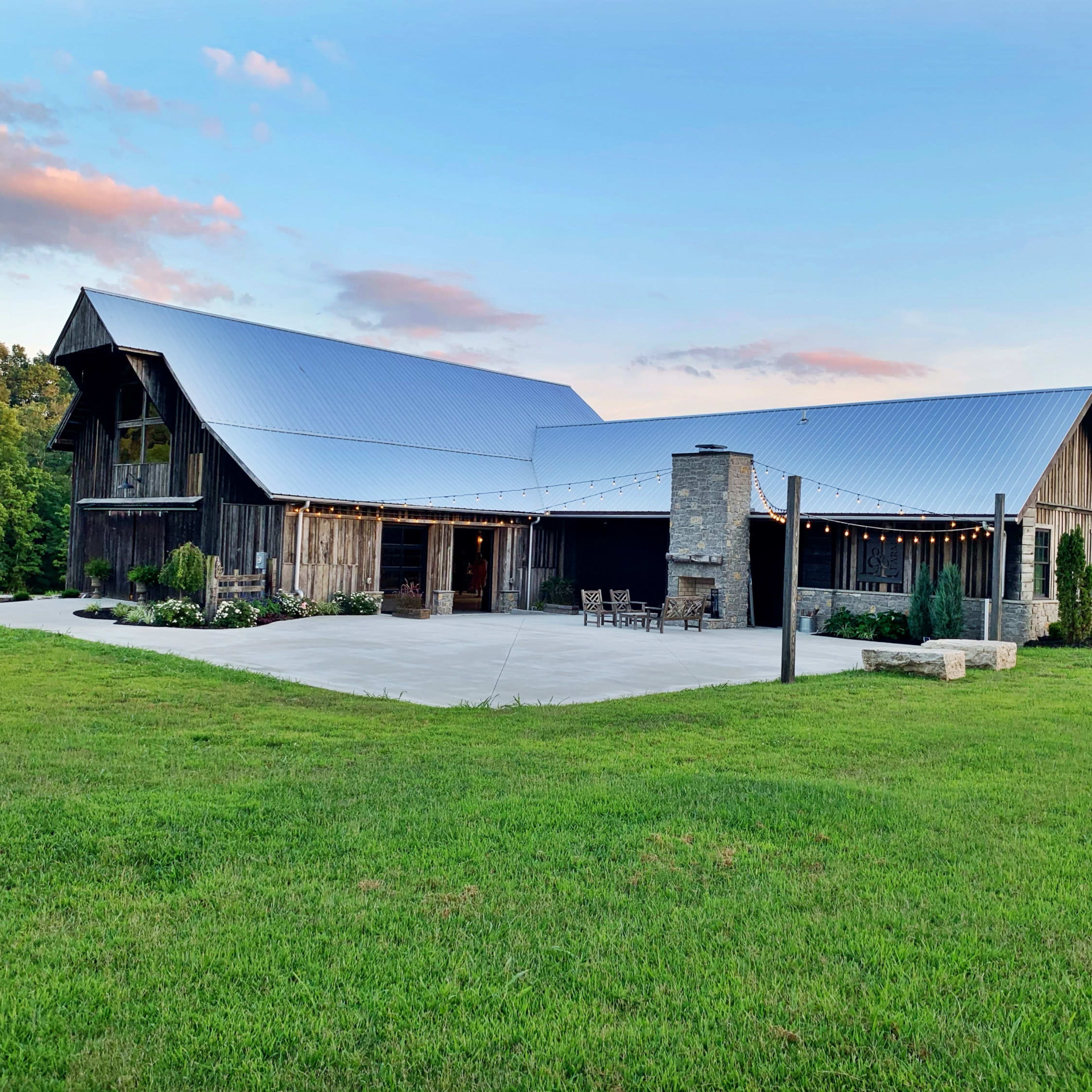 A large, modern barn-style building with a metal roof is set on a grassy landscape, featuring a stone fireplace and outdoor seating area.