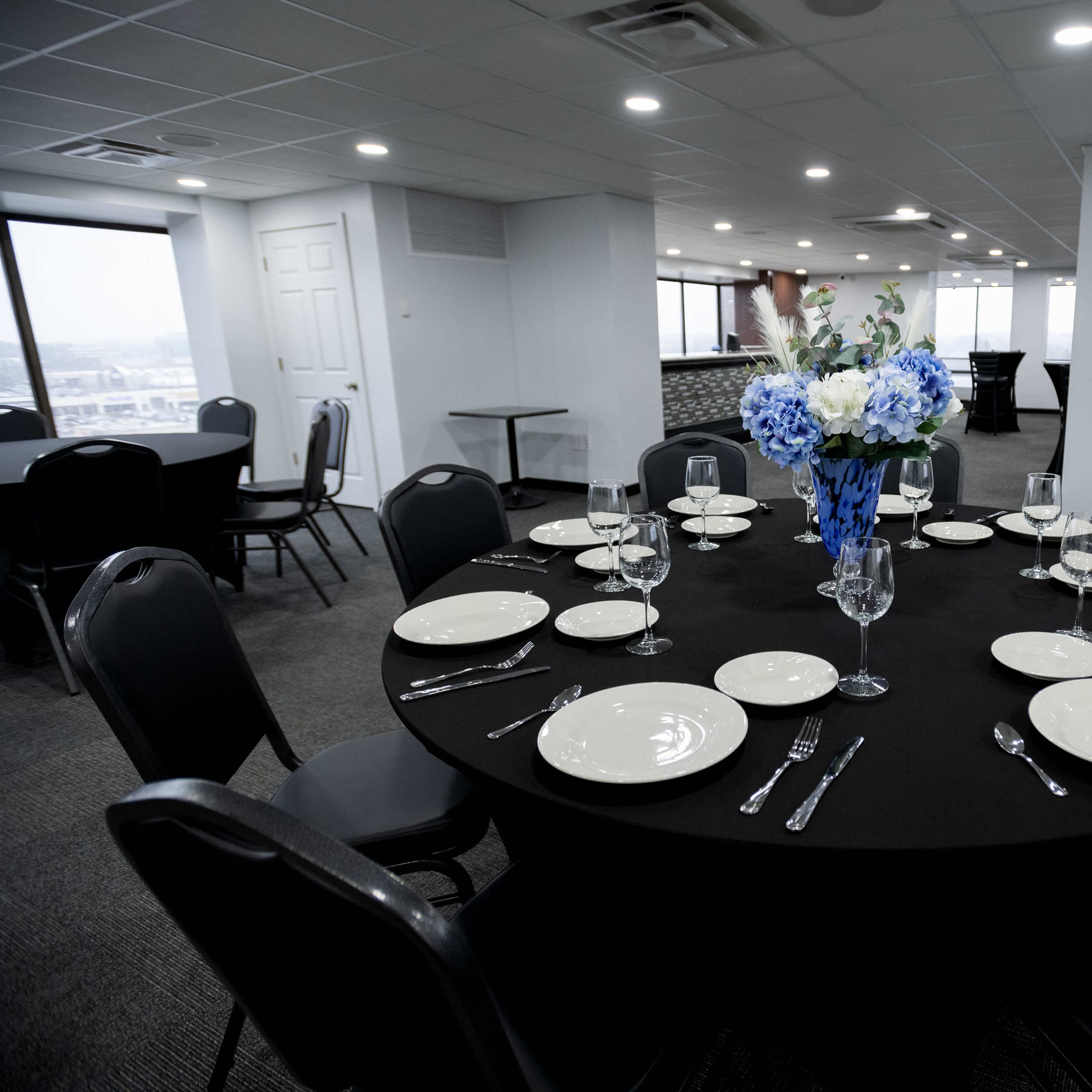 A round table set with white plates, glassware, and silverware is decorated with a blue floral centerpiece in a well-lit event space.