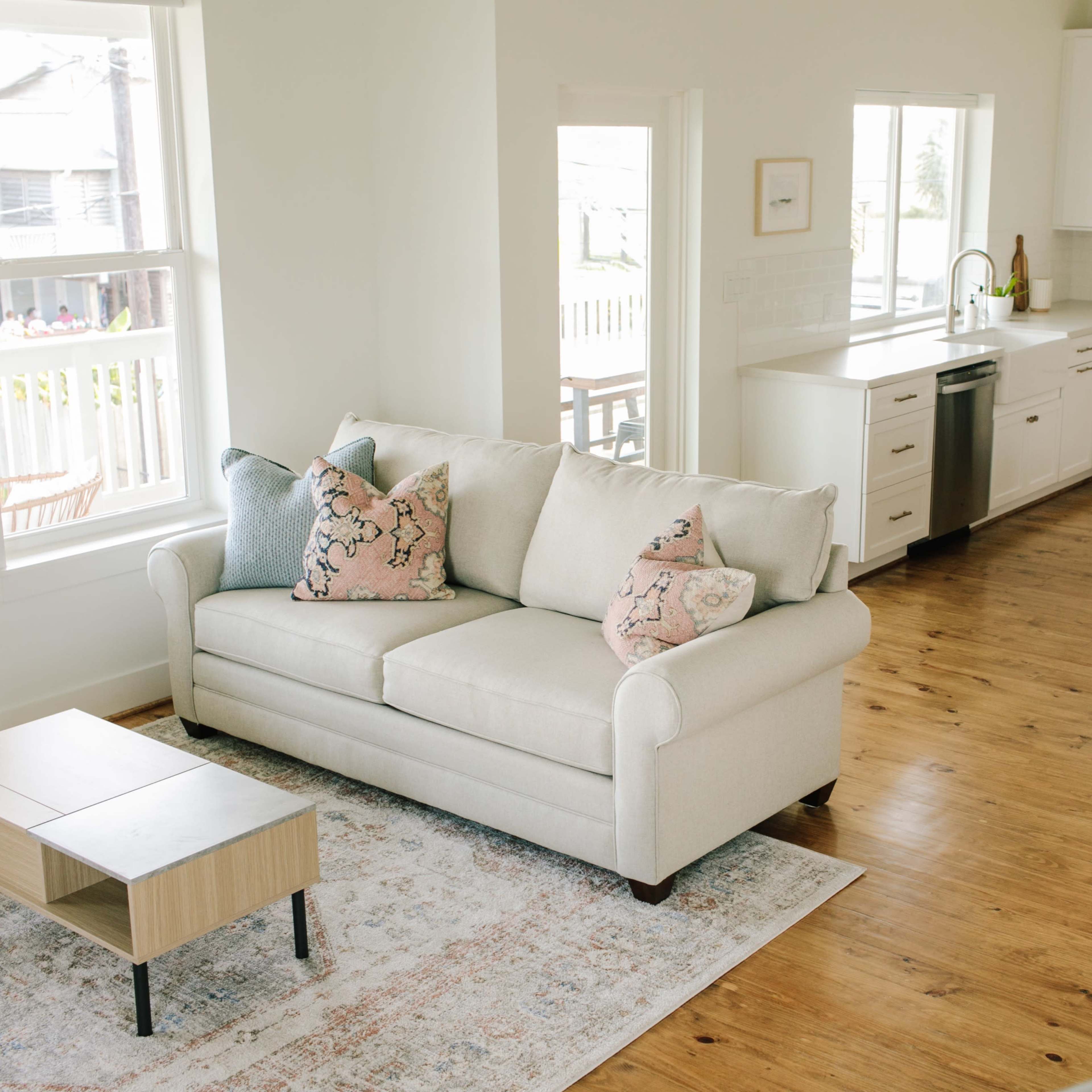 A light-filled open living area featuring a beige couch with decorative pillows, a wooden coffee table, and a kitchen area with white cabinets and barstools.