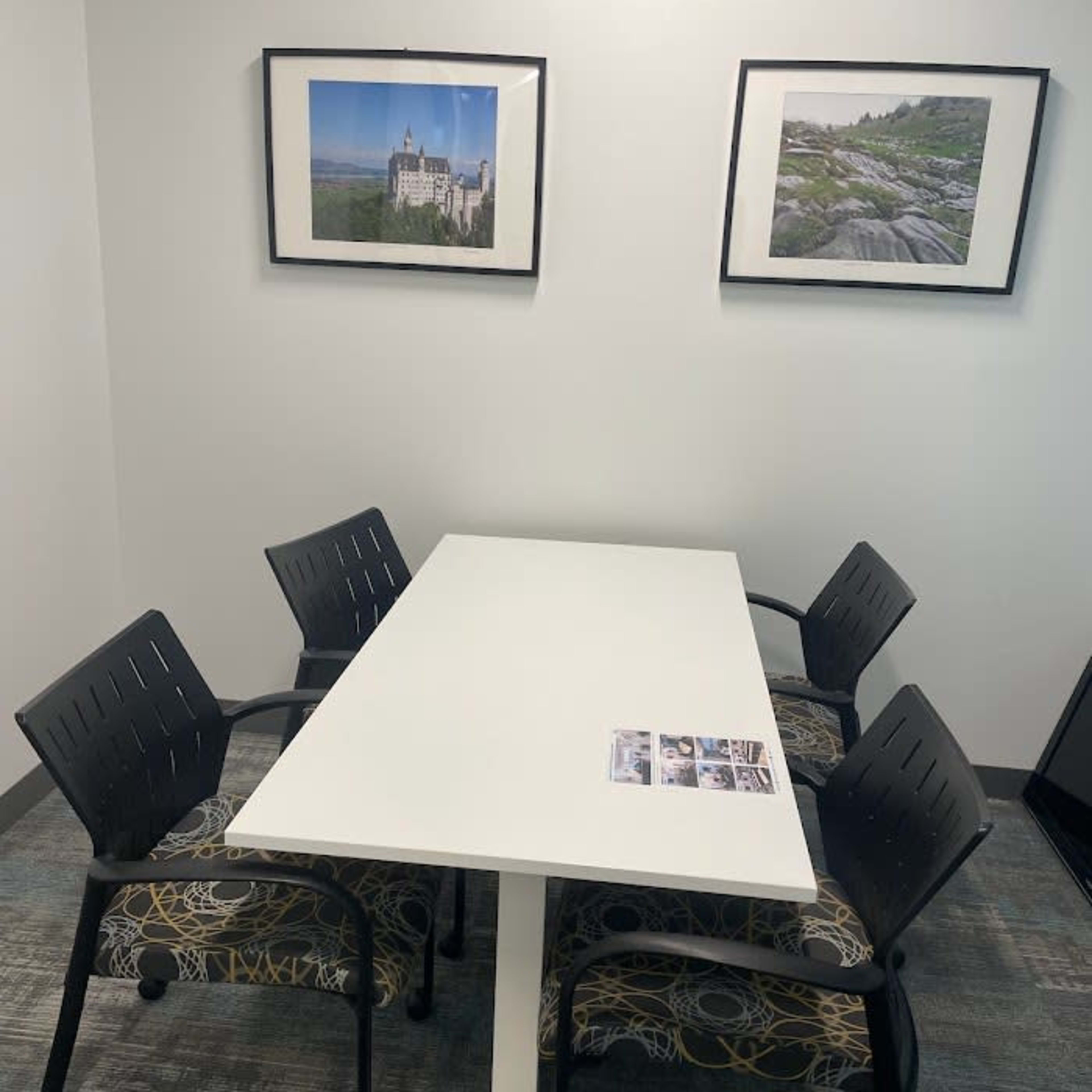 A small meeting room with a white table and four black chairs, featuring two framed photographs on the walls and a brochure on the table.