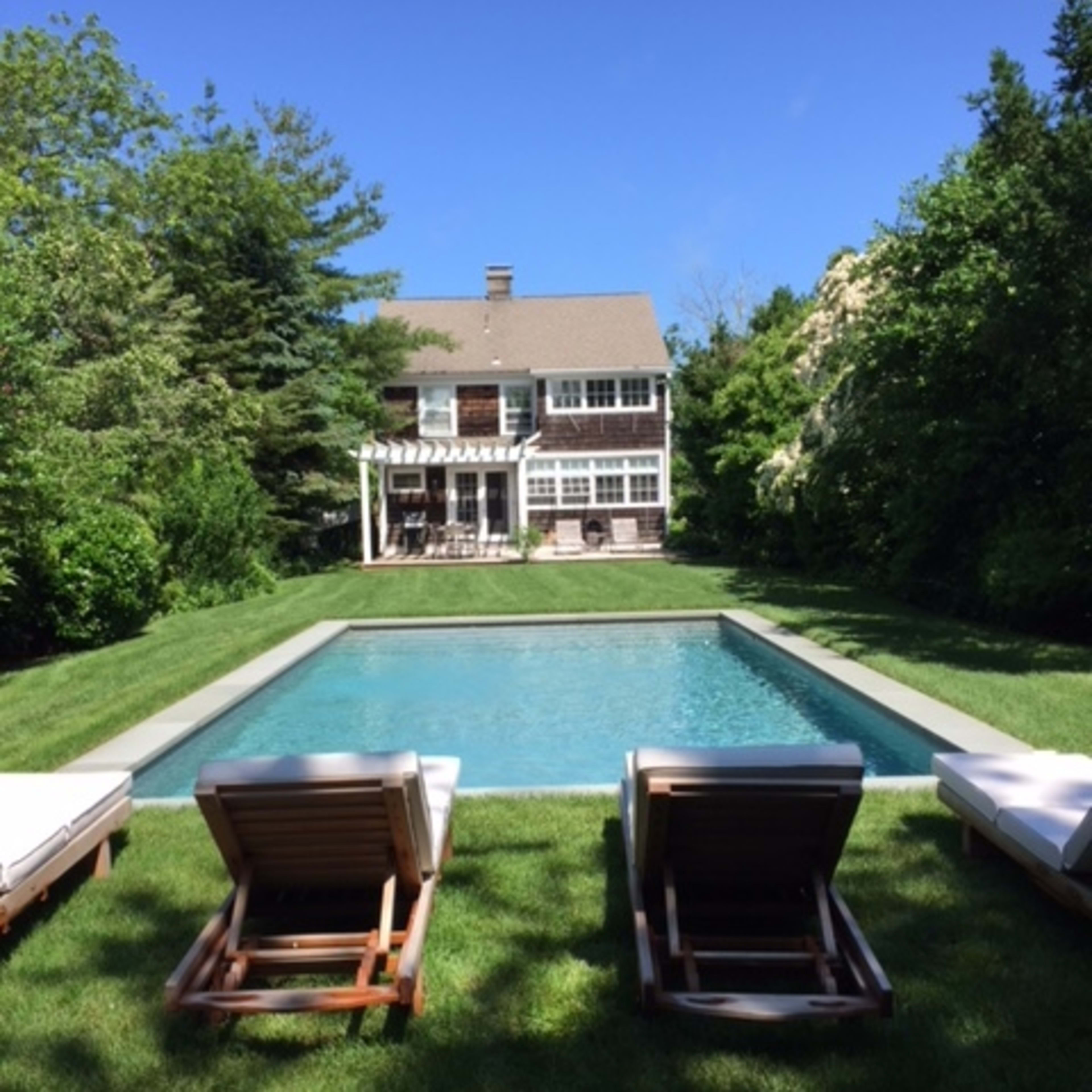 A modern house with a green lawn and a rectangular swimming pool is surrounded by trees and shrubs.