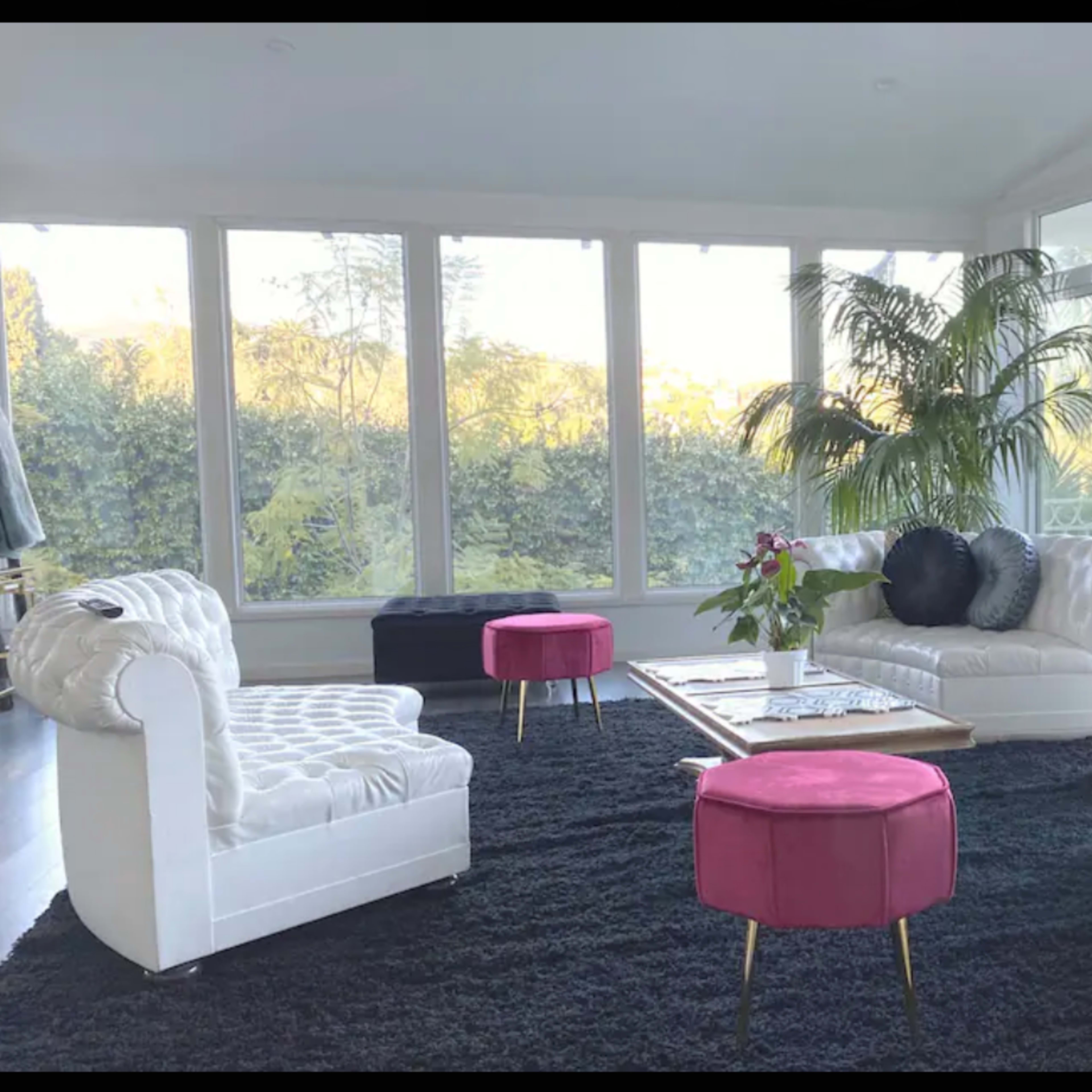 A modern living room features white tufted sofas, a pink ottoman, and large windows overlooking greenery.