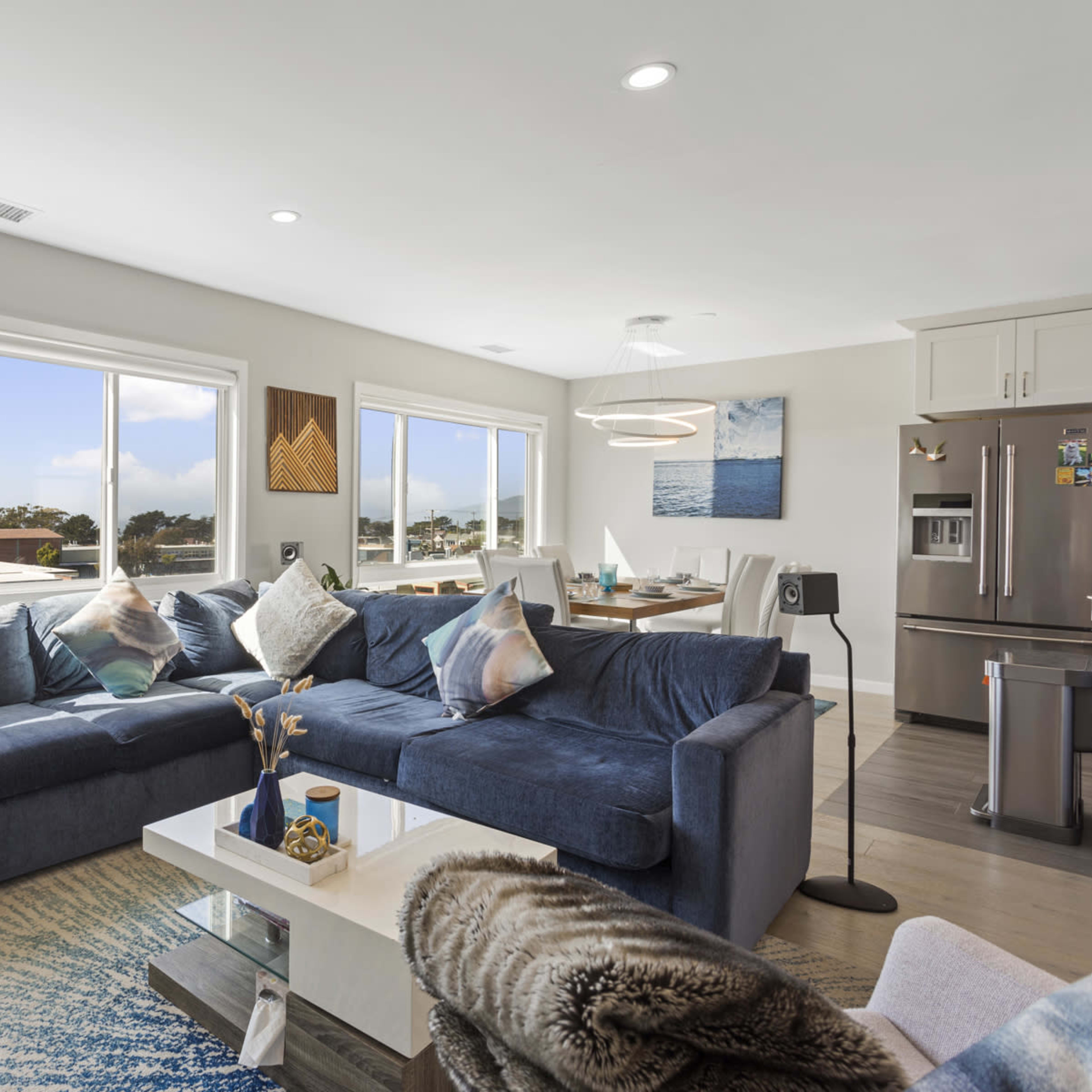 A modern living room with large windows, a blue sectional sofa, and a dining area set with a table and chairs.