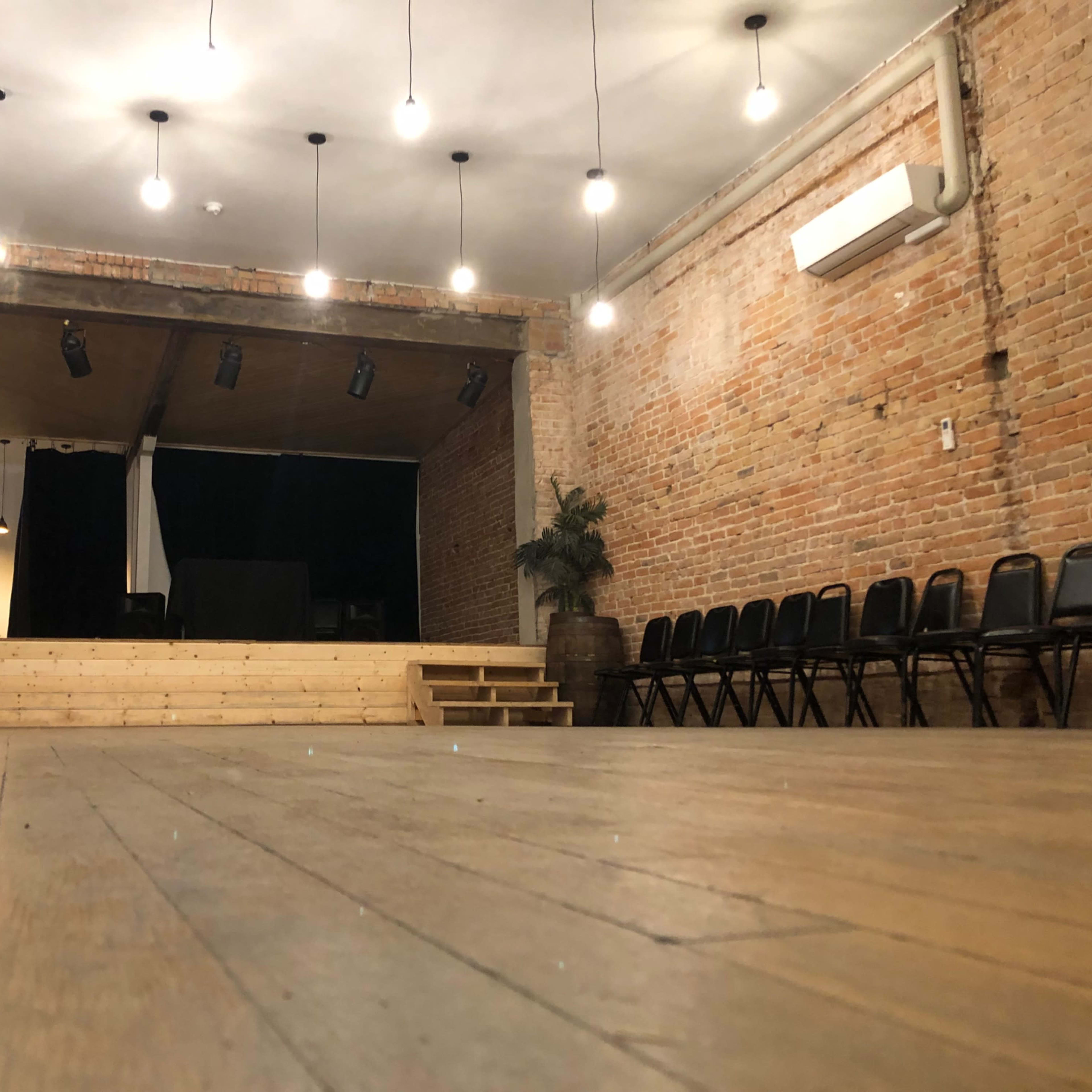 A wooden stage with steps is illuminated by hanging lights in a brick-walled performance space, with black chairs arranged in rows nearby.