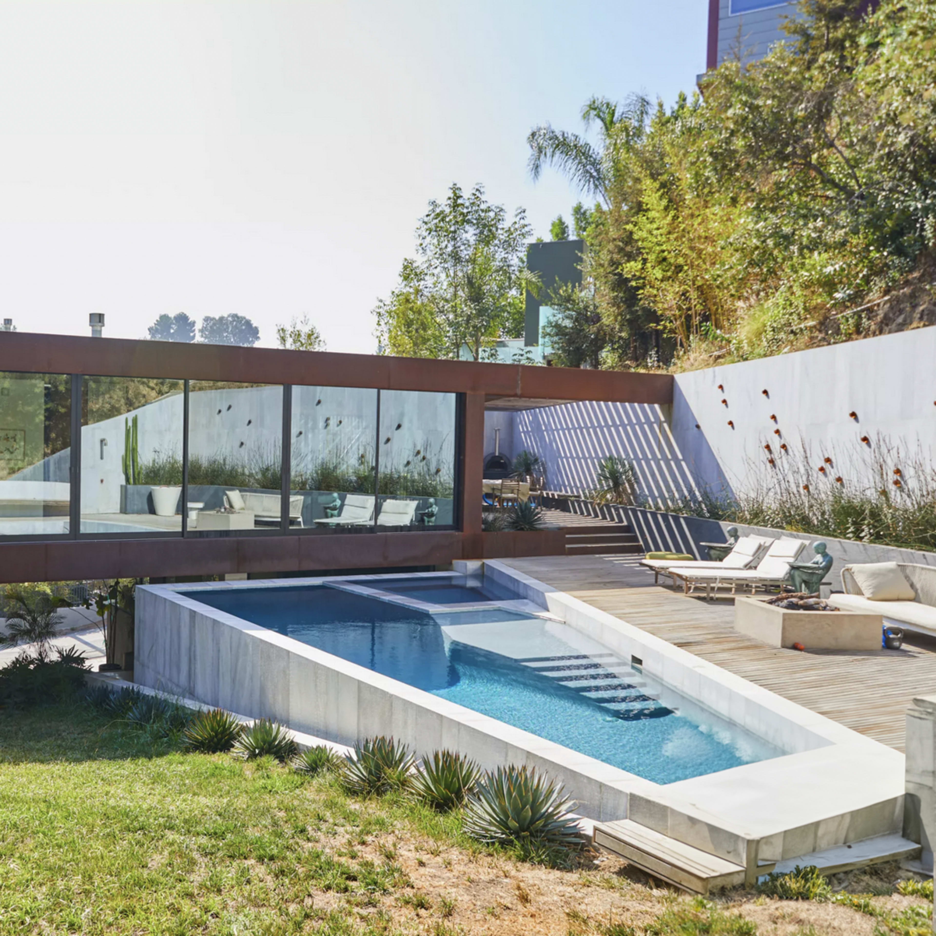 A modern house with large glass windows overlooks a rectangular swimming pool surrounded by landscaped greenery.
