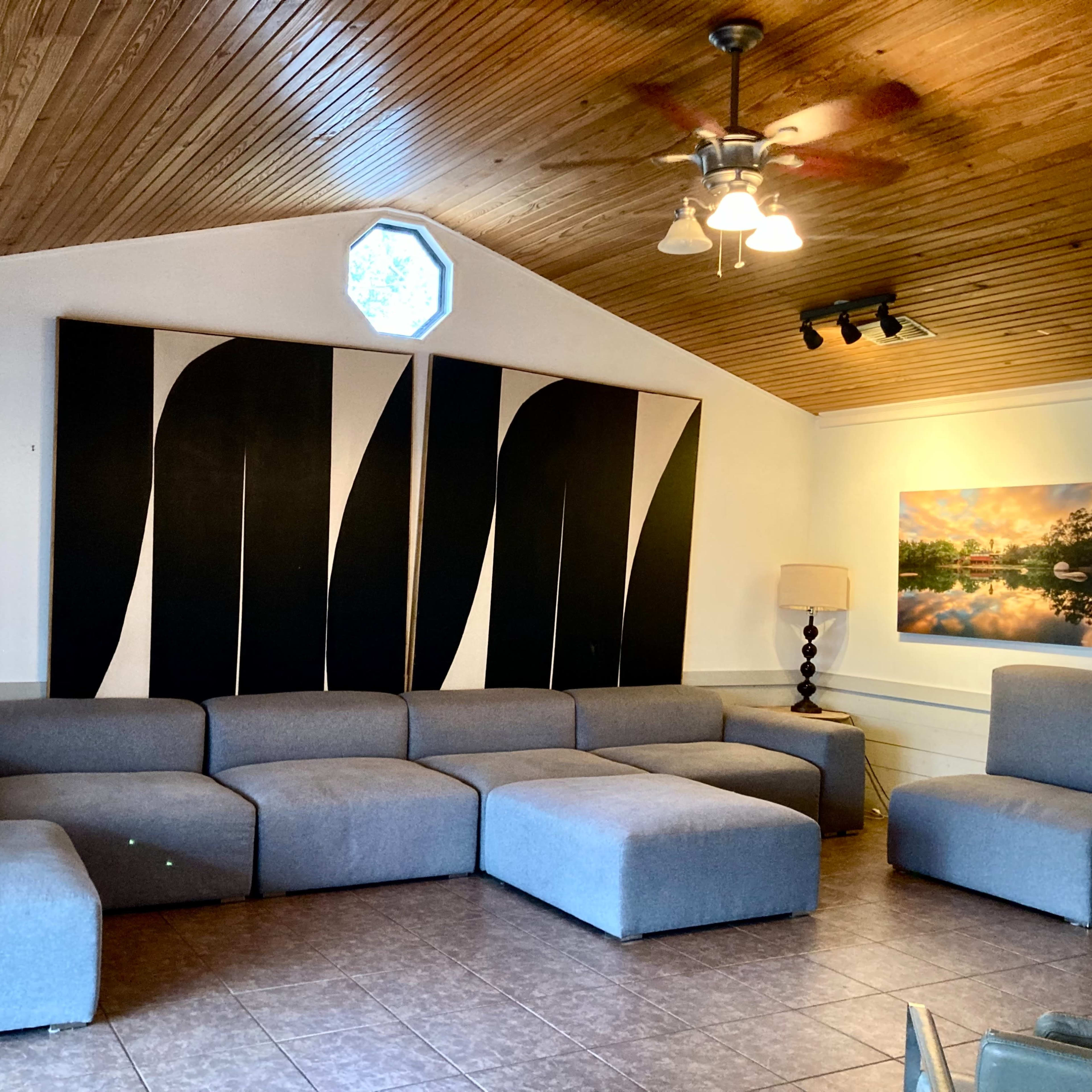 A spacious living room features a large gray sectional sofa, a wall-mounted lamp, and two pieces of wall art alongside a window with a unique octagonal shape.